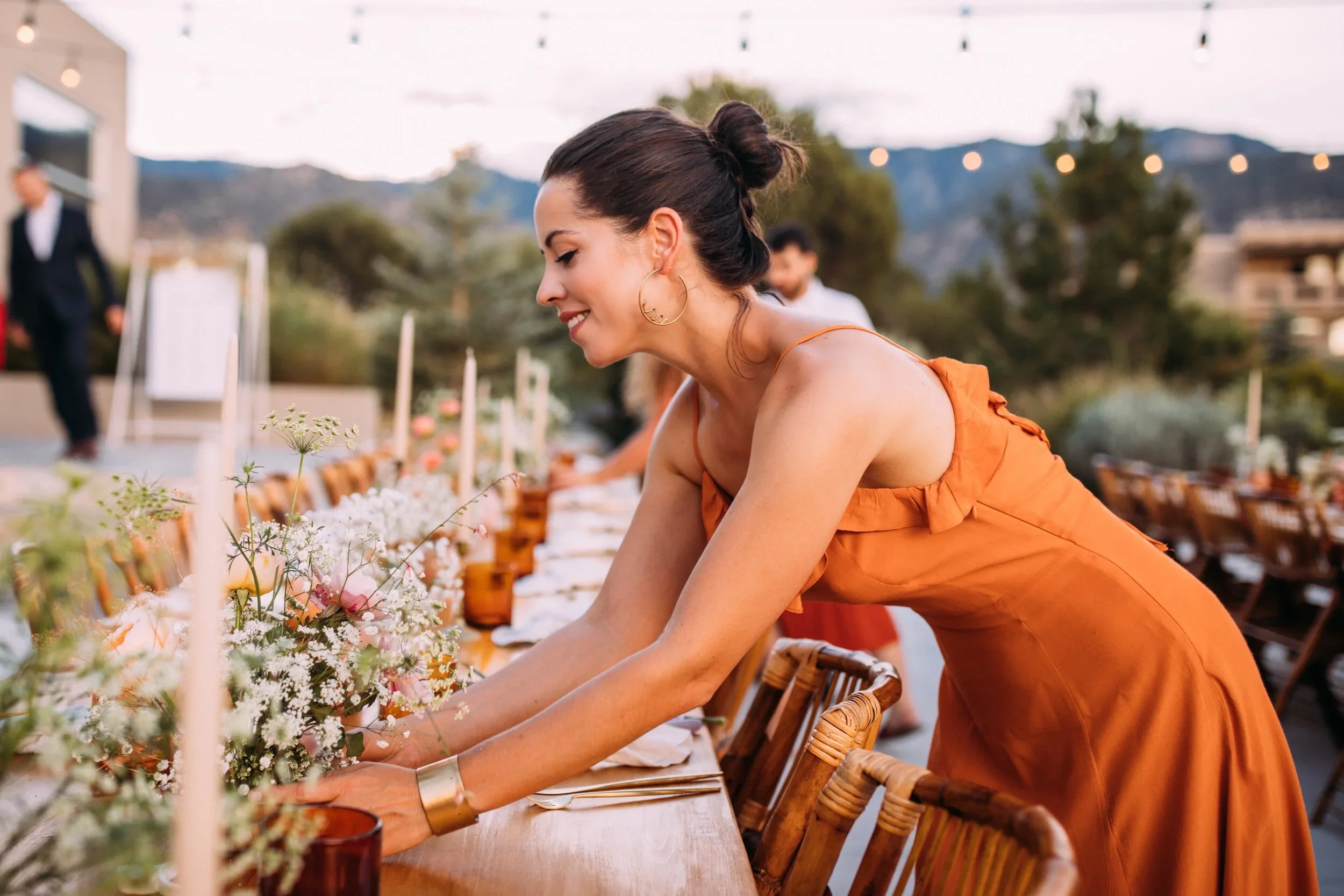 Woman in an orange dress arranging flowers on a long table at an outdoor event during sunset with string lights overhead and mountains in the background.