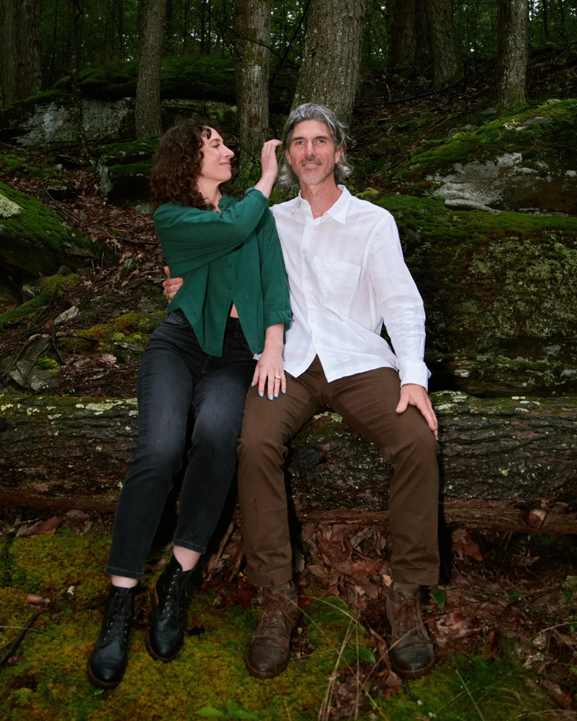 A woman and a man sitting on a fallen tree trunk in a forest, surrounded by trees, moss, and rocks, with the woman touching the man's hair.