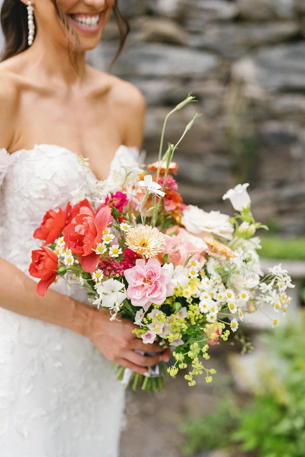 A bride in a white lace wedding dress holding a large bouquet of colorful flowers, smiling, with a stone wall in the background.