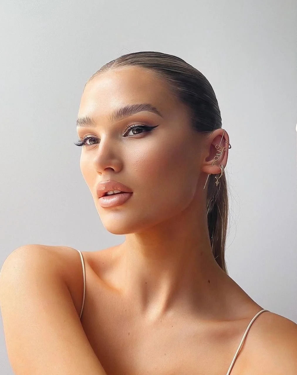 A woman with slicked-back hair and multiple ear piercings, wearing makeup with winged eyeliner, poses against a plain background.