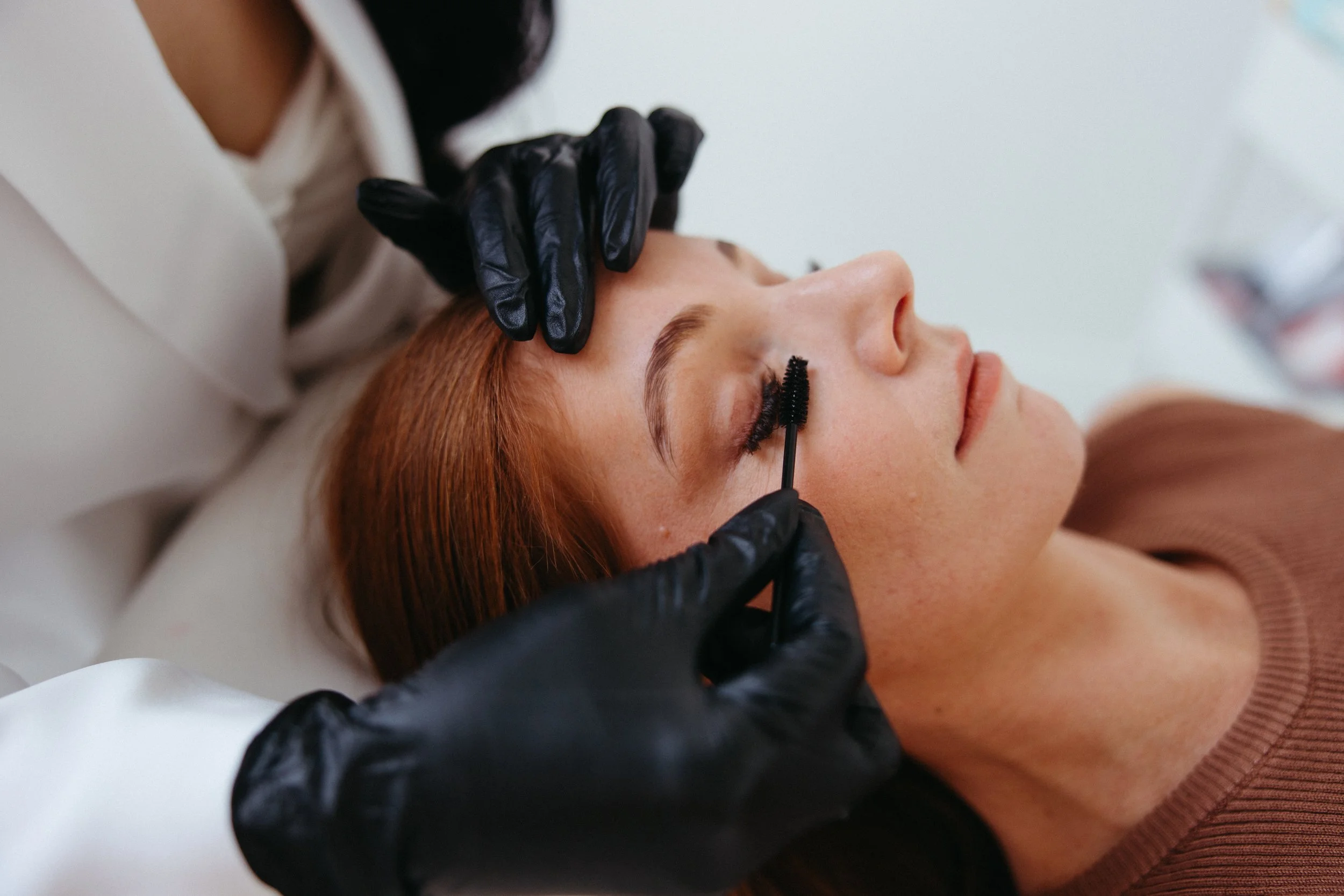 Person applying mascara to a woman's eyelashes using a spoolie brush, while wearing black gloves.