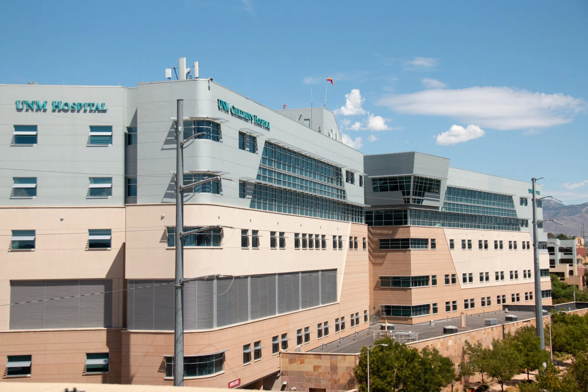 A large multi-story hospital building with parking lots filled with cars in the foreground, surrounded by trees and other buildings in an urban area.