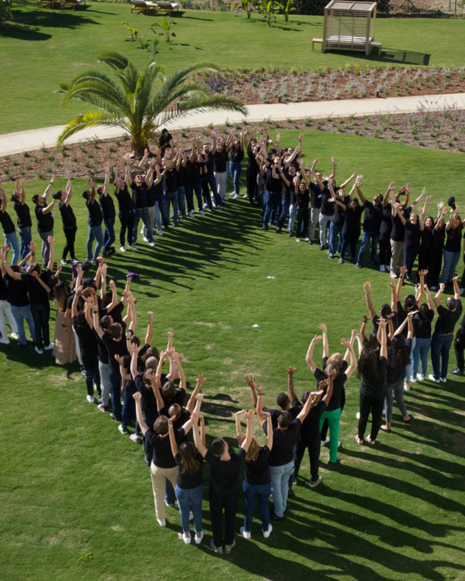 A large group of people standing in a heart shape on a grassy lawn, raising their arms in celebration, with a palm tree and landscaped area in the background.