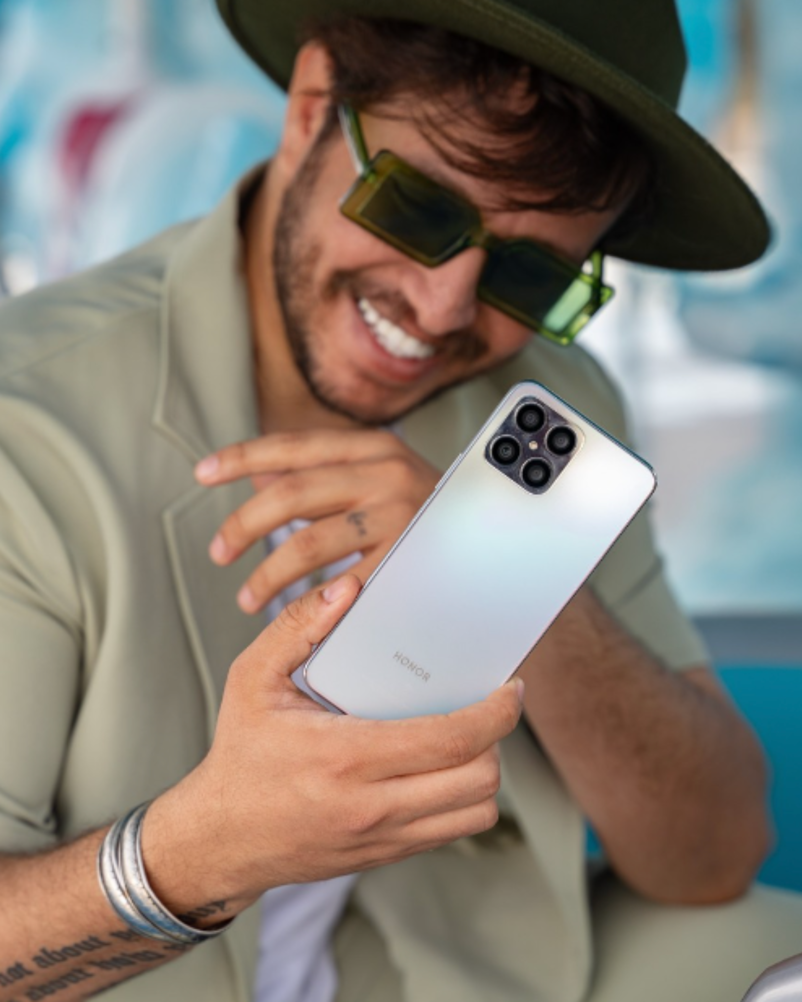 A man smiling and looking at a white Honor smartphone, wearing sunglasses, a hat, and a light-colored shirt.
