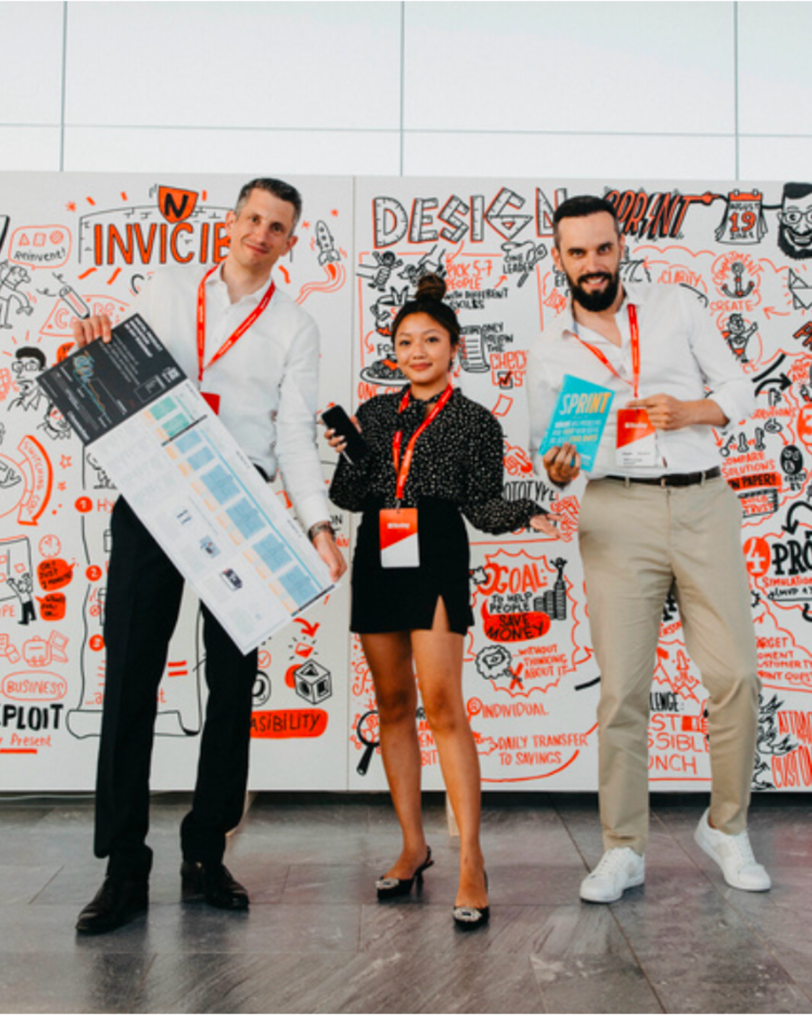 Three diverse people standing in front of a wall covered with business-related drawings and words at a conference. They hold conference materials and wear lanyards with name tags.