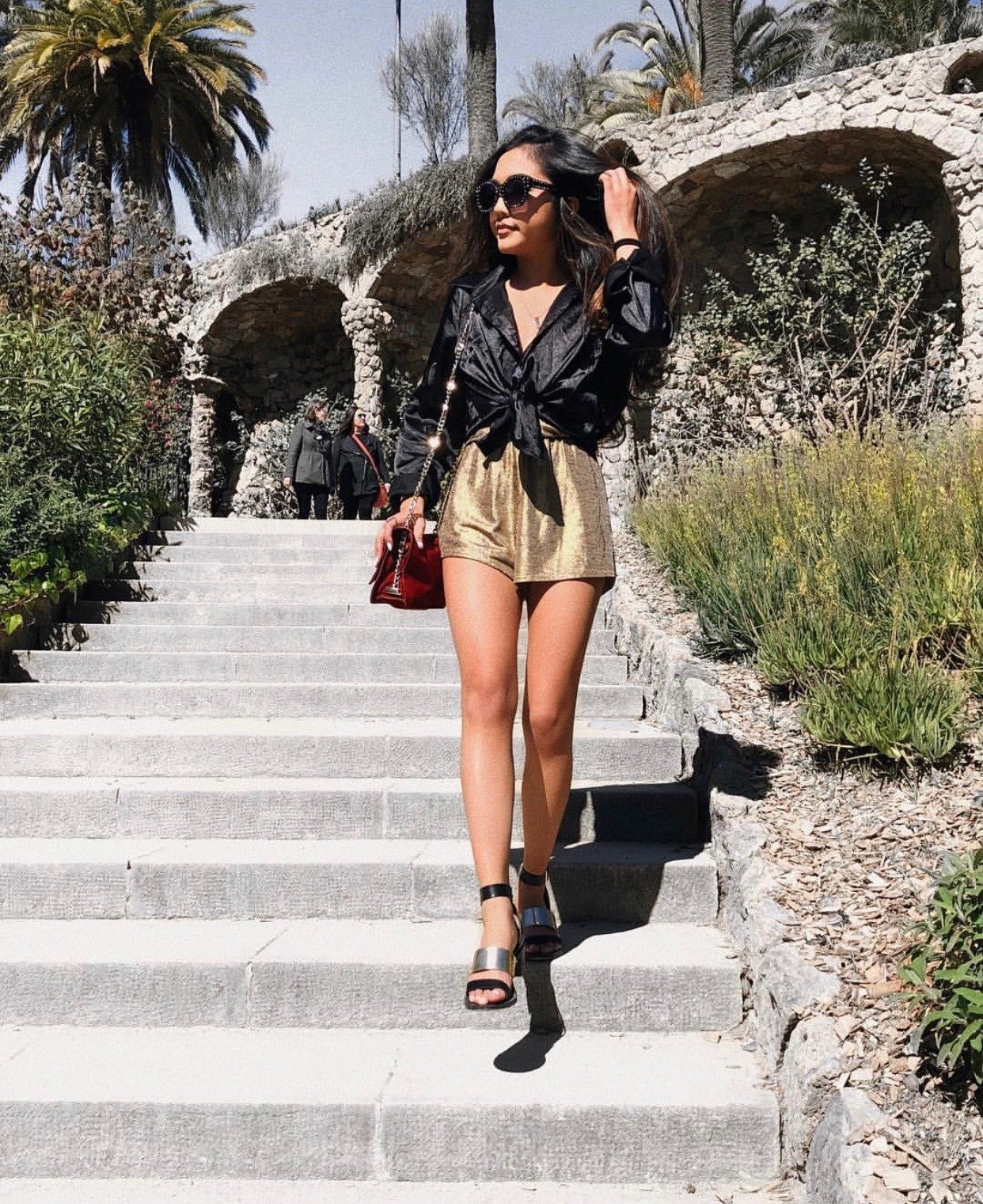A woman walking down outdoor steps in a sunny, landscaped area with palm trees and stone architecture, wearing a black jacket, gold shorts, and high heels, carrying a red purse and wearing large sunglasses.