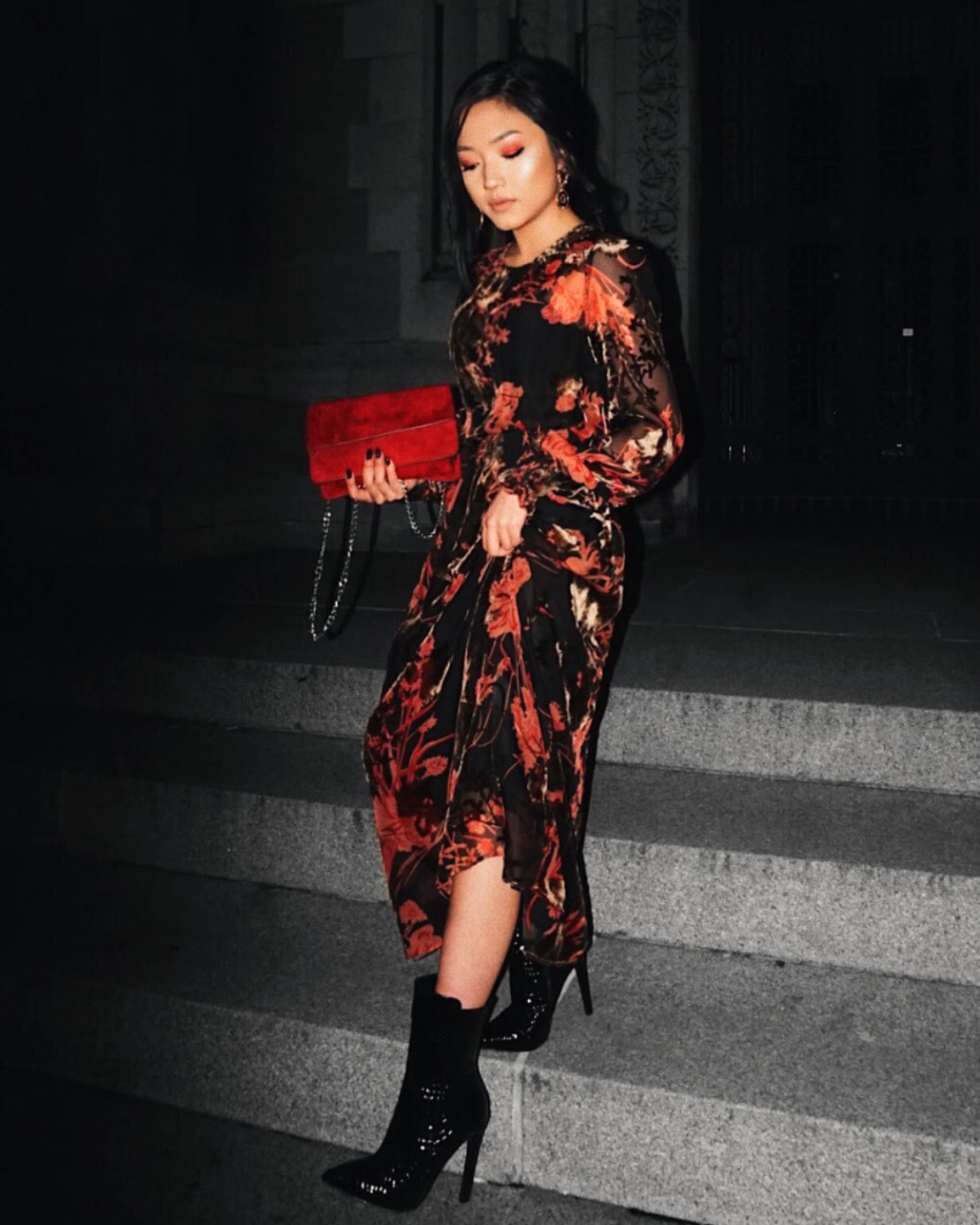A woman in a black and red floral dress with high heels, holding a red clutch, standing on steps at night.