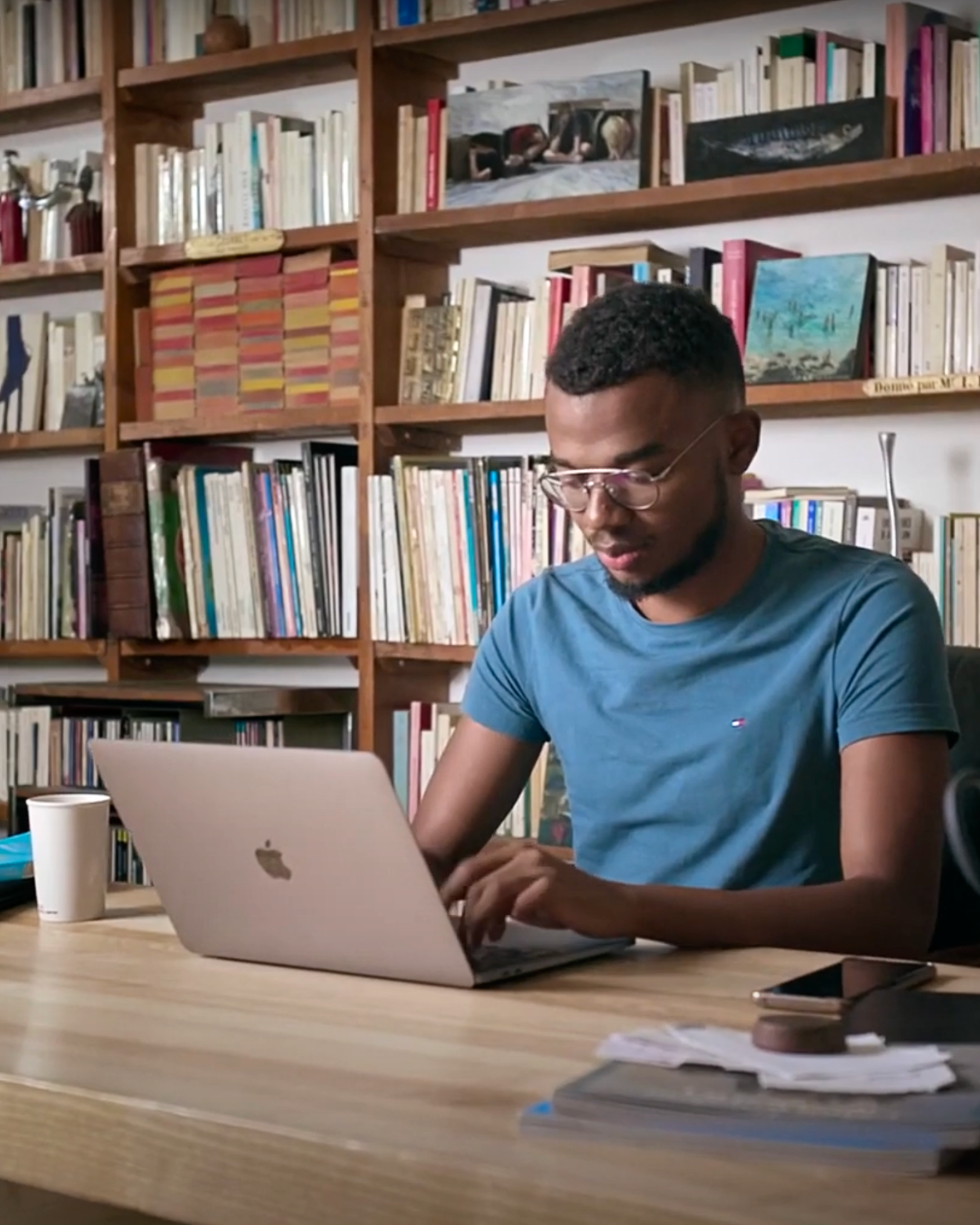 A man in glasses and a blue t-shirt working on a silver MacBook in a room with wooden bookshelves filled with books and paintings.