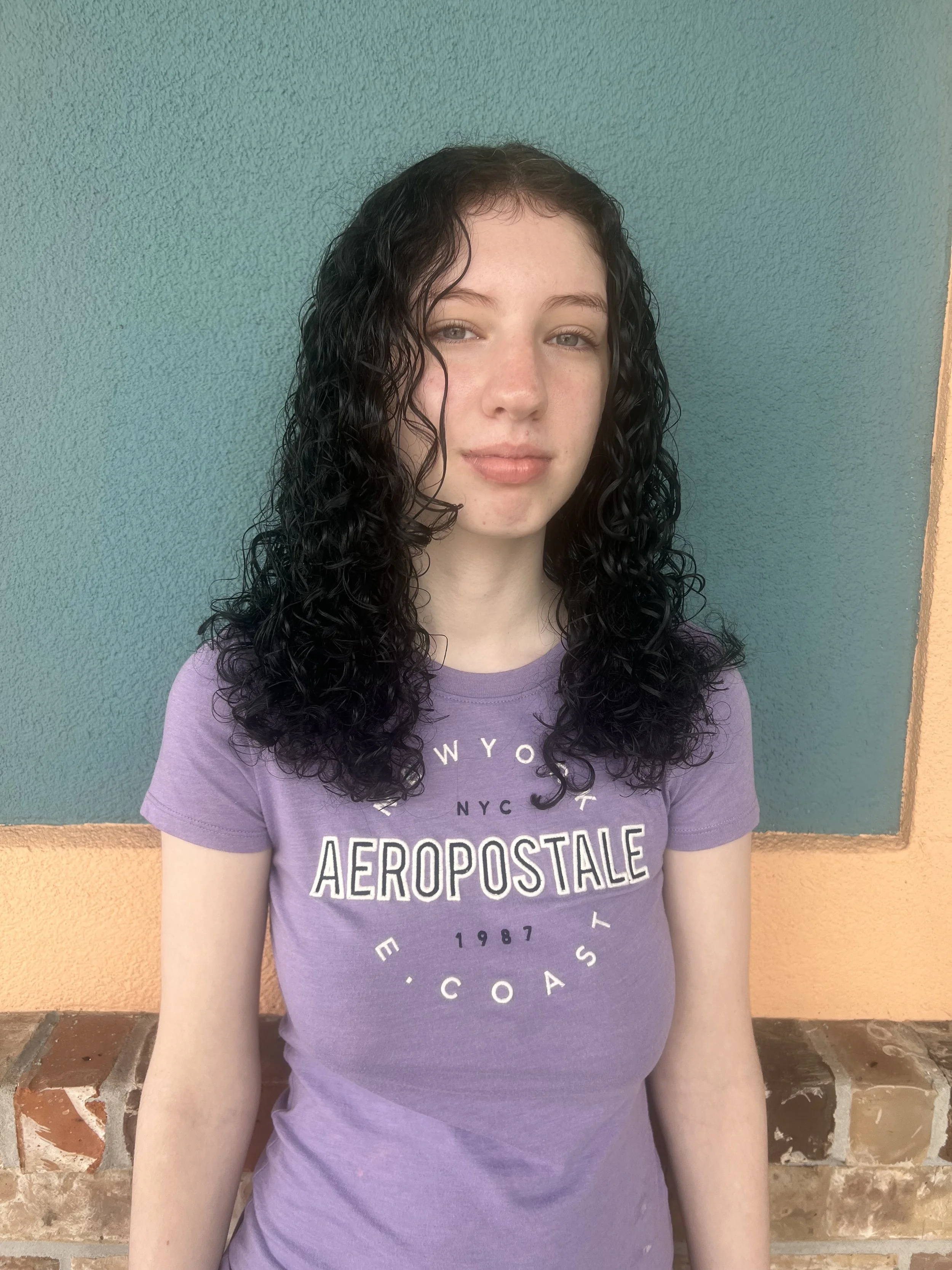 Young woman with curly black hair standing against a colorful wall, wearing a purple Aeropostale t-shirt.