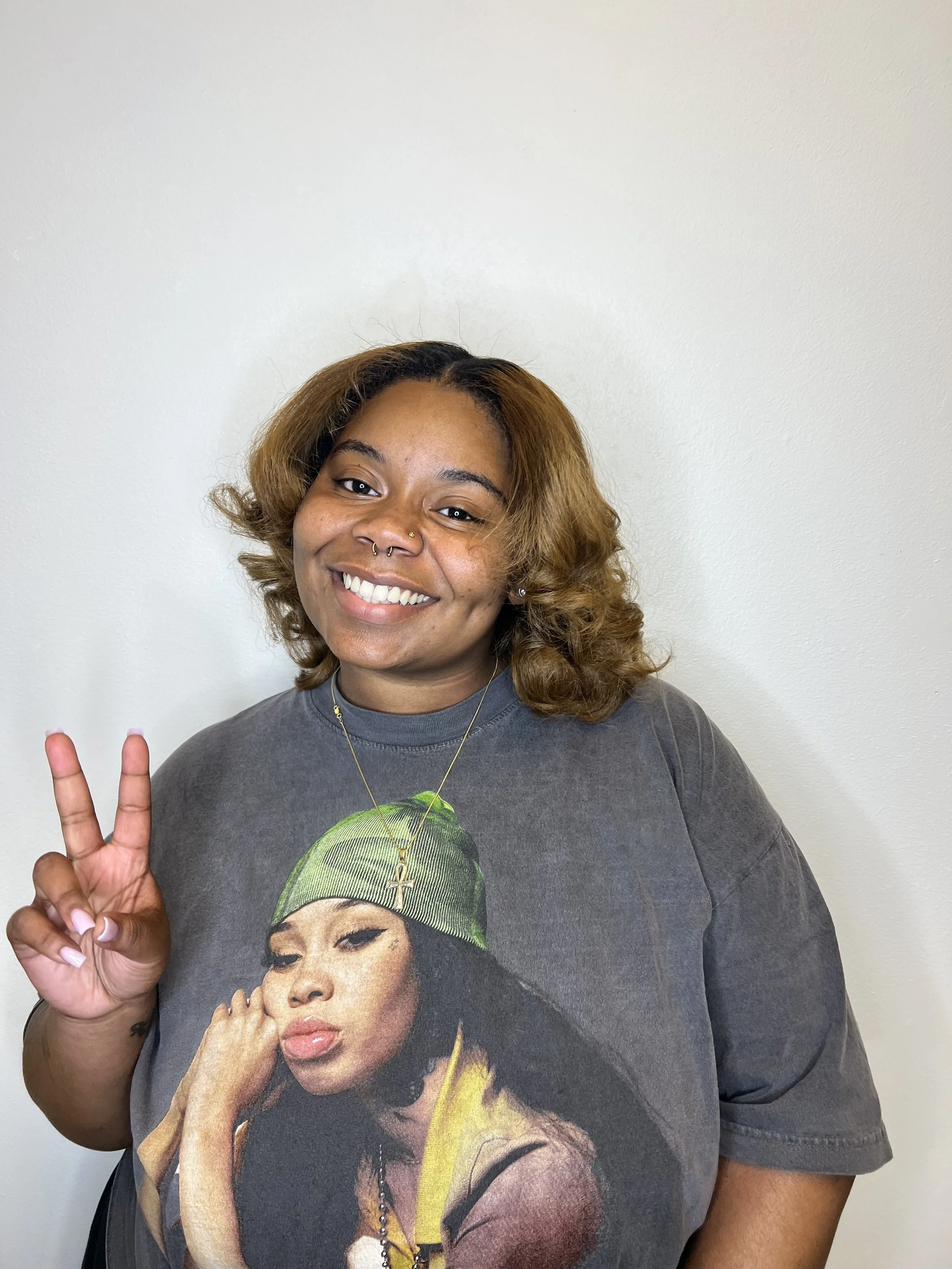 A young woman with shoulder-length hair, smiling, making a peace sign, wearing a graphic T-shirt featuring a woman with a green bandana.