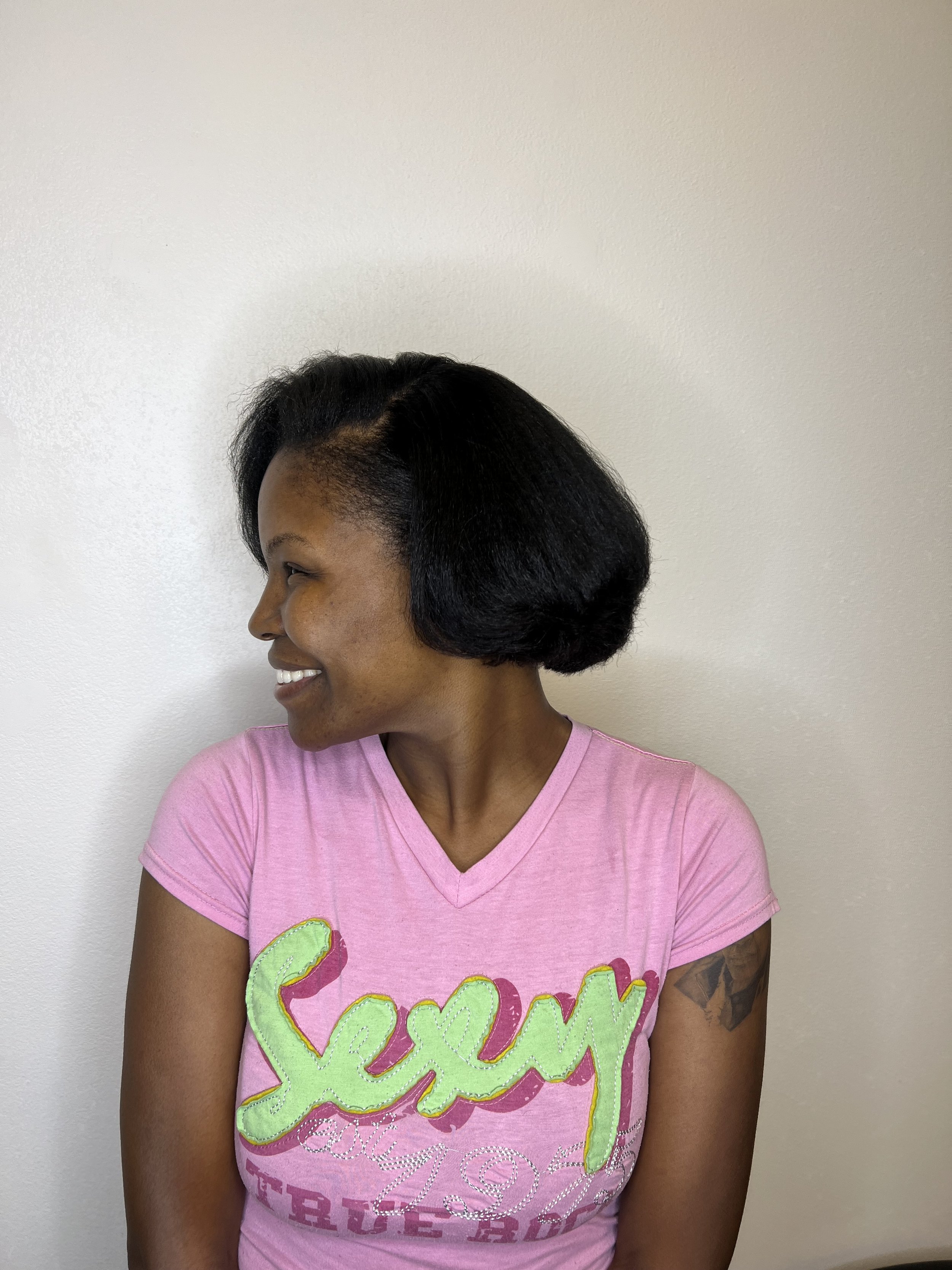 A woman with black hair styled in a bob, wearing a pink T-shirt with colorful cursive writing, smiling and looking to her left against a plain wall.