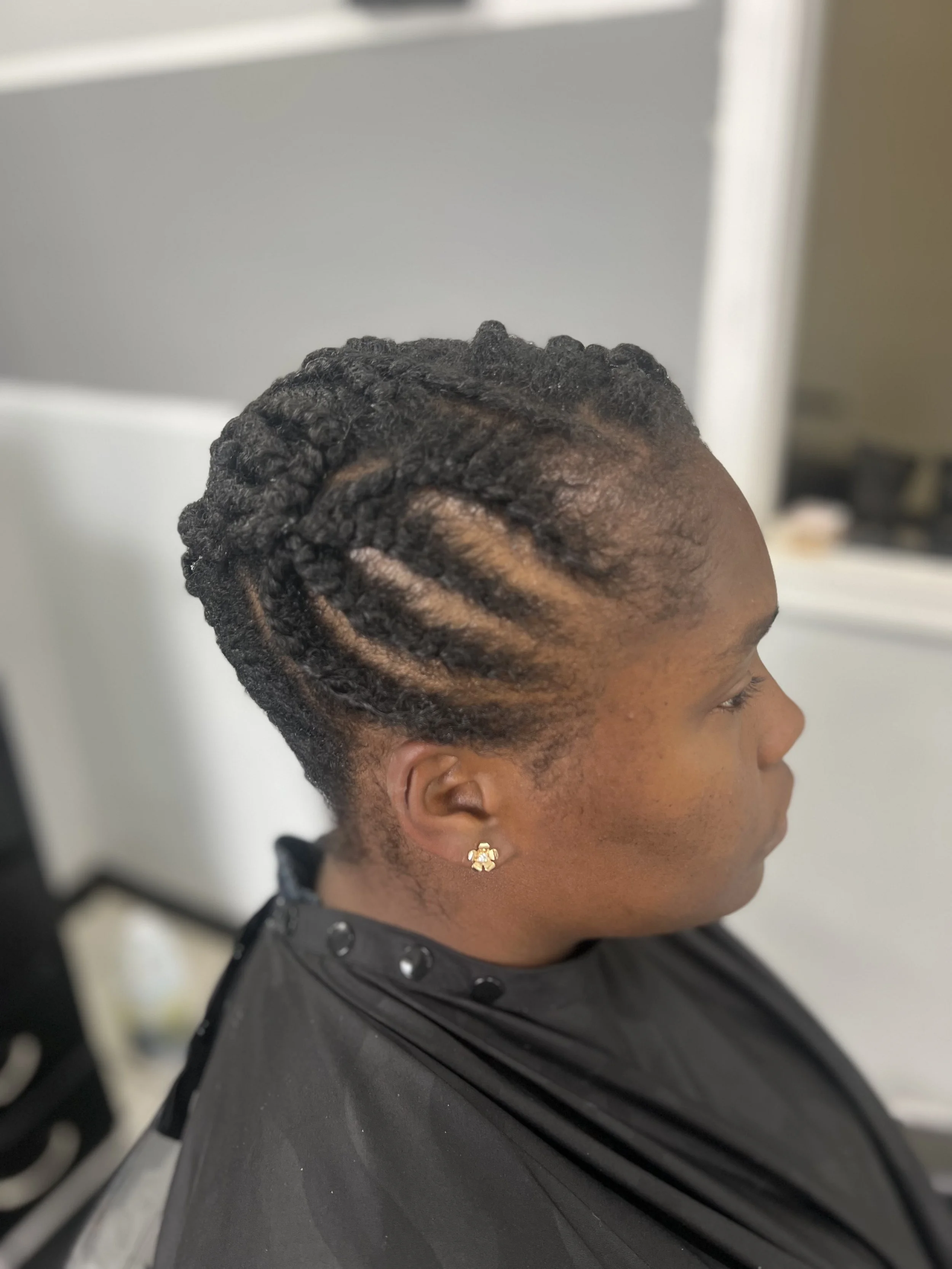 A woman with a short hairstyle of neatly twisted natural hair in tight, defined twists, wearing a black cape in a salon.