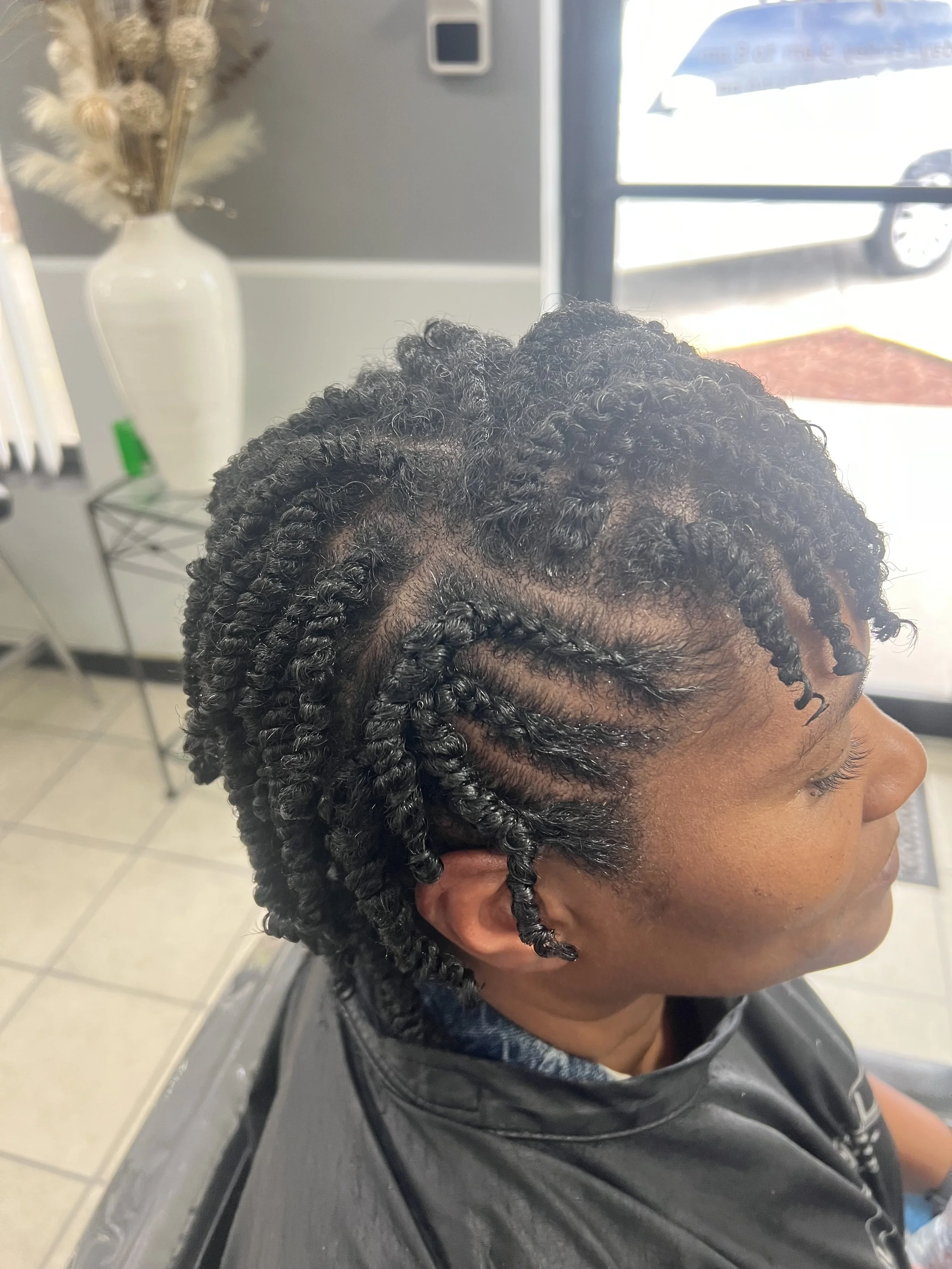 Side view of a person with freshly styled black, tight, curly twists in their hair, sitting in a salon chair. The background shows a window, a white vase with dried flowers, and a small table with salon supplies.