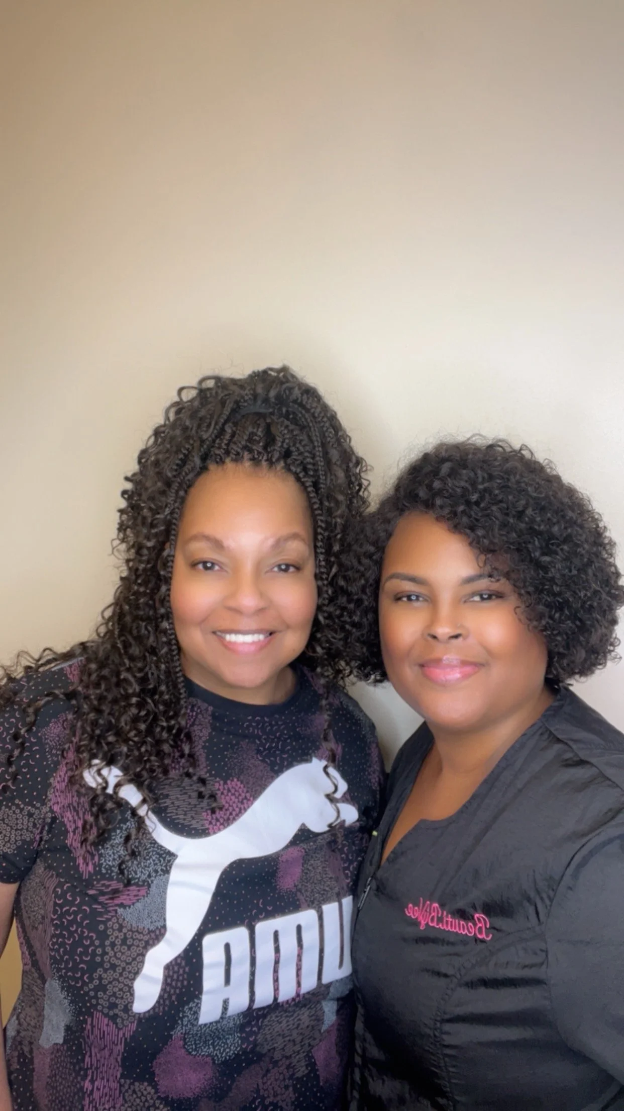 Two women with curly hair smiling, standing close together against a plain beige wall.