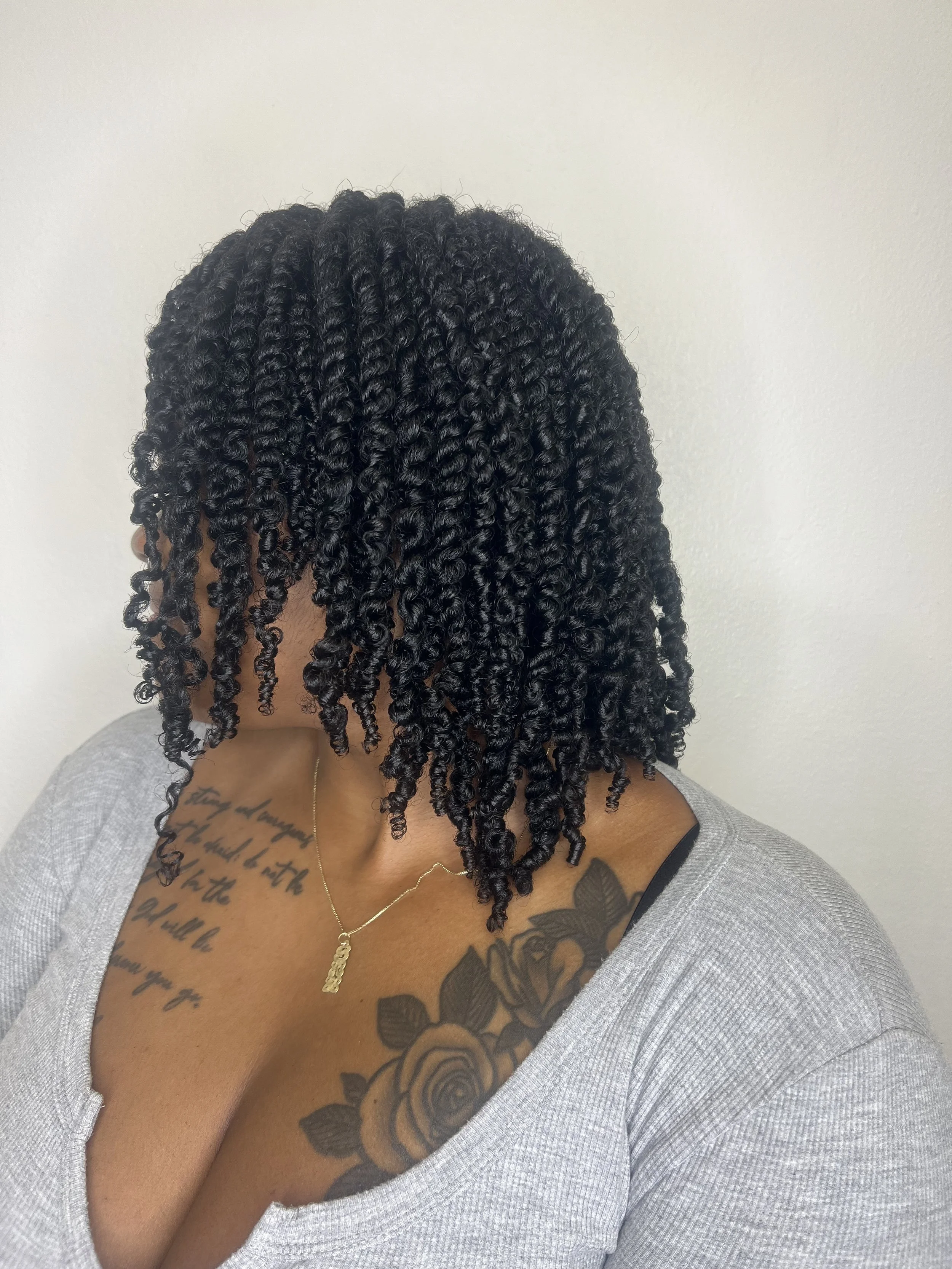 Woman with shoulder-length, curly black hair wearing a gray top and a gold necklace with a pendant, showing tattoos on her chest and shoulder, against a plain white wall.