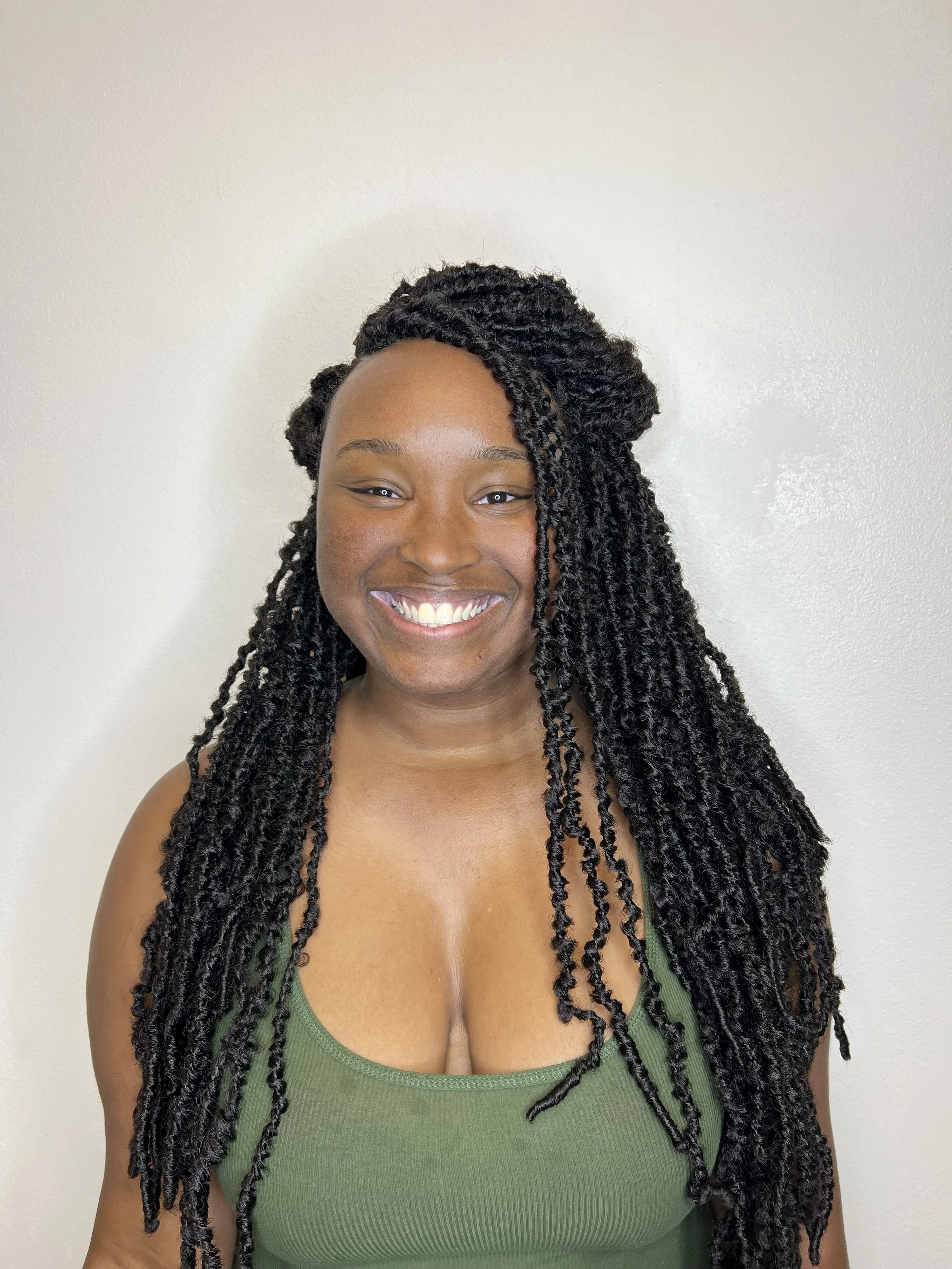 A smiling woman with long twisted black hair wearing a green top against a light-colored wall.