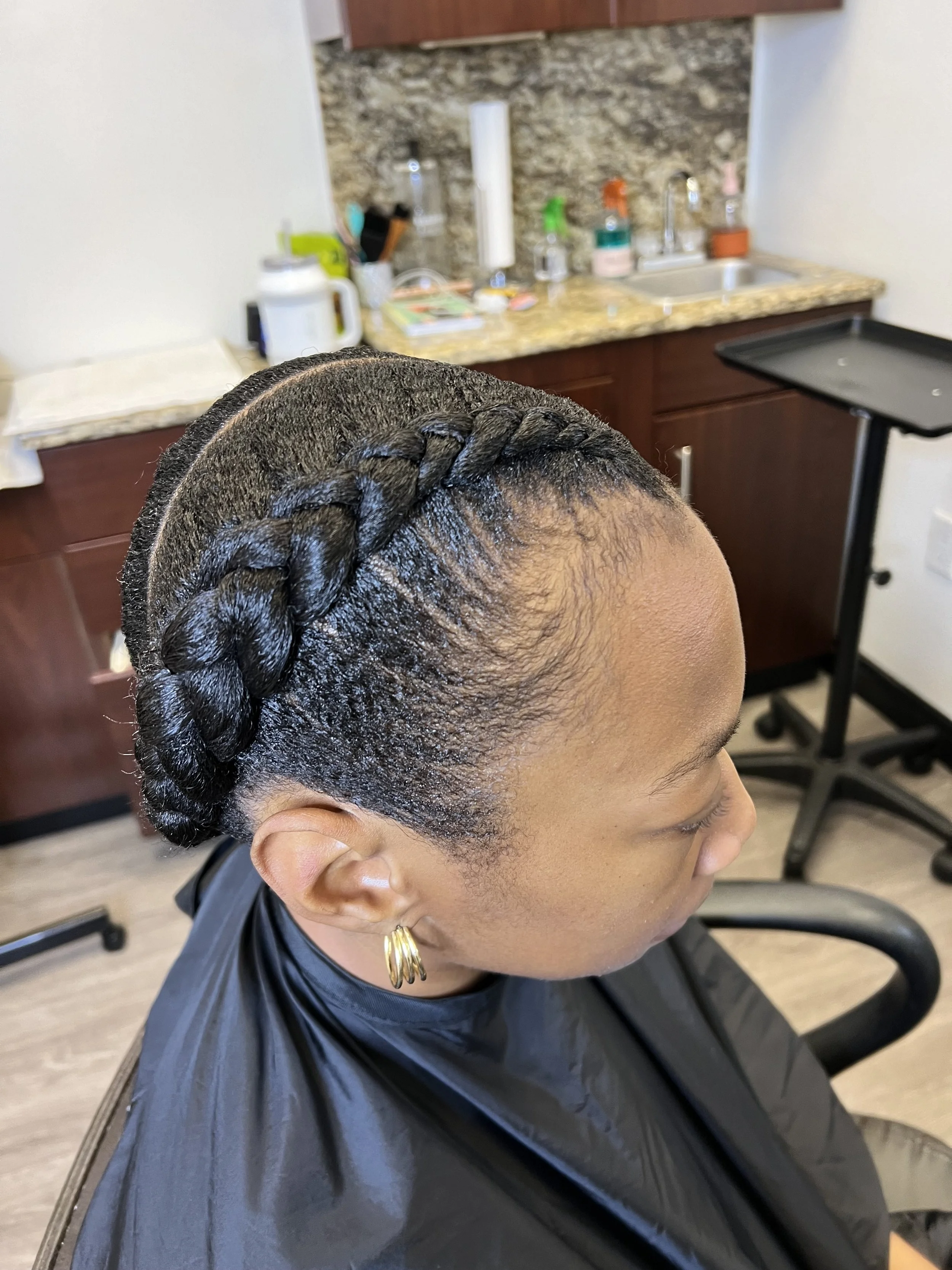 A woman sitting in a salon chair with her hair styled in a neat braided updo, wearing gold hoop earrings, in a salon with a cherry wood cabinet and marble countertop.