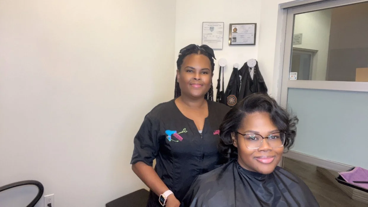 A hairstylist standing behind a woman with styled hair and glasses inside a salon.