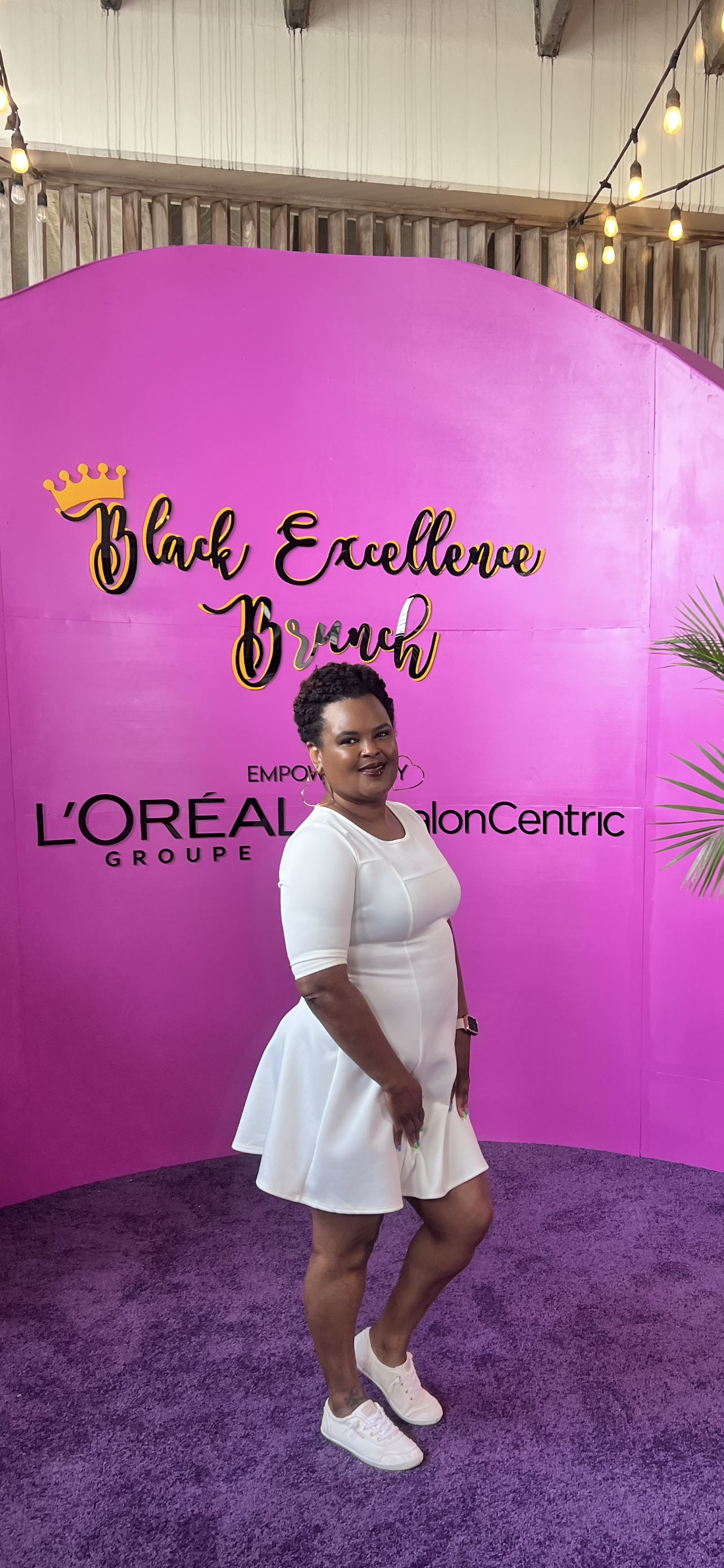 A woman in a white dress standing in front of a pink backdrop with event branding, with string lights overhead and a purple carpet underfoot.