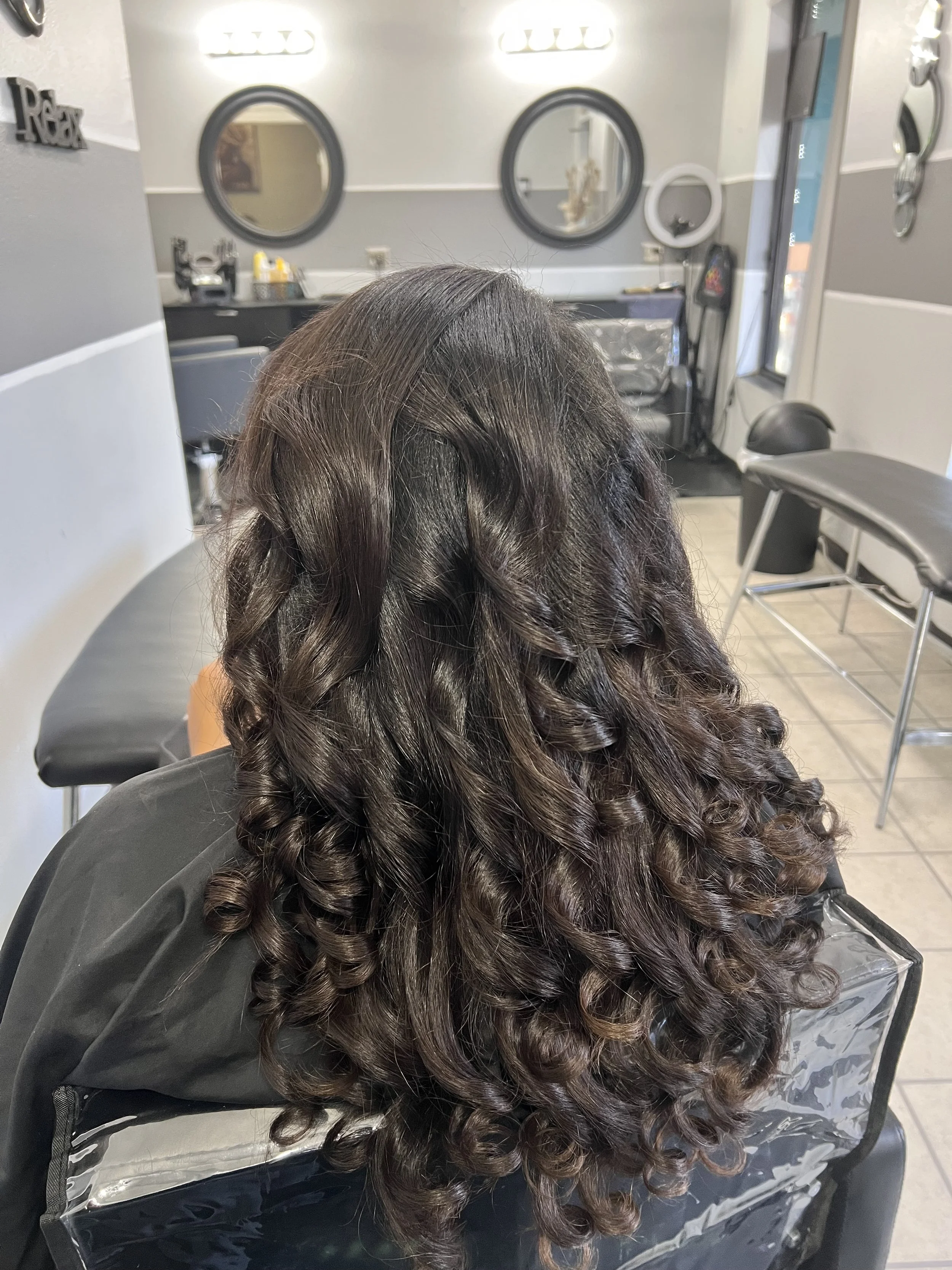 Hair stylist chair with long, dark, curly hair in a salon setting, mirrors and salon equipment visible in the background.