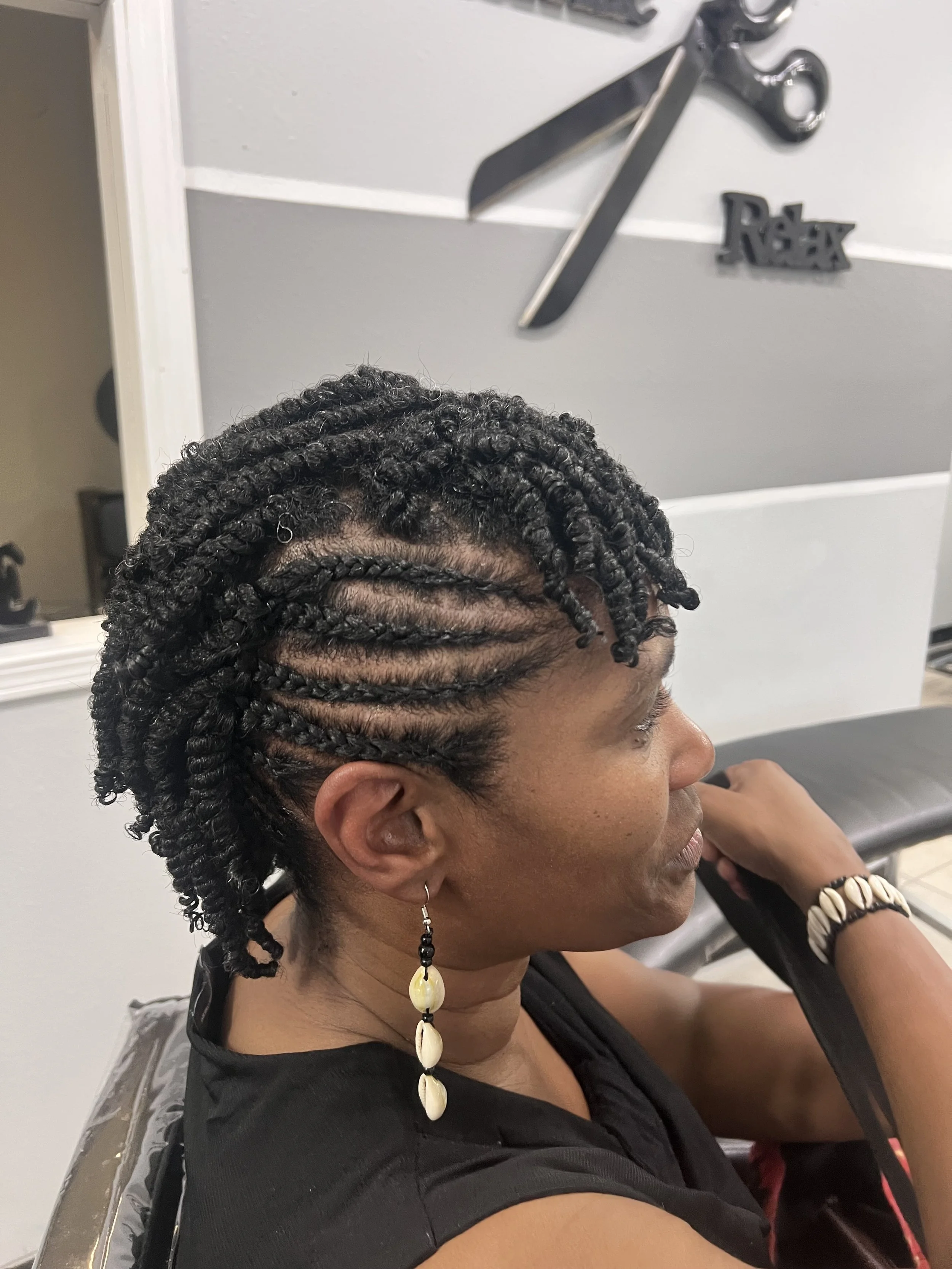 Side view of a woman with black curly twist hairstyle, wearing shell earrings and a black shirt, sitting in a hair salon with a mirror and hairdressing tools in the background.