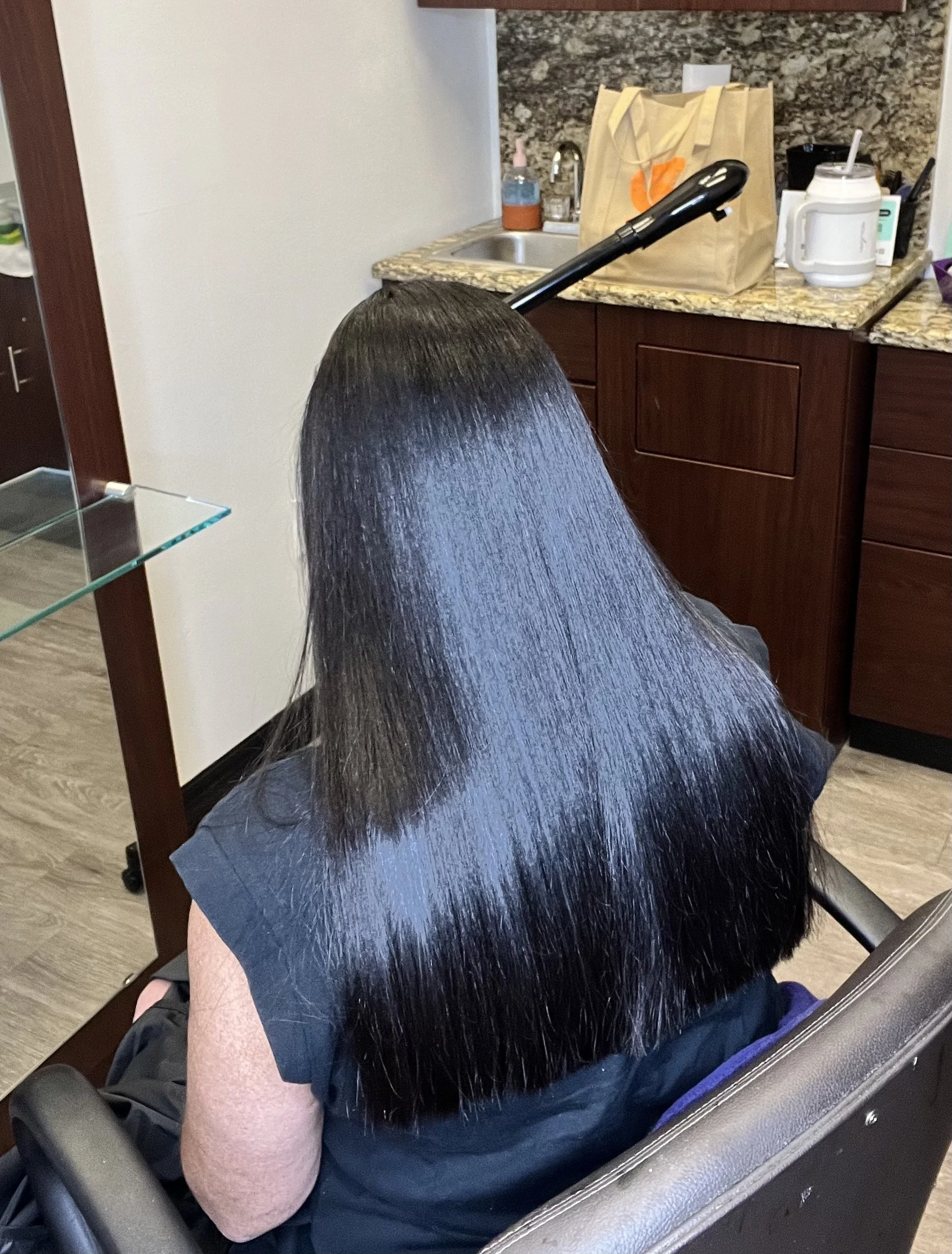 Person with long, straight, shiny black hair sitting in a salon chair, facing away from the camera, in front of a hair styling station with a countertop and kitchen sink.