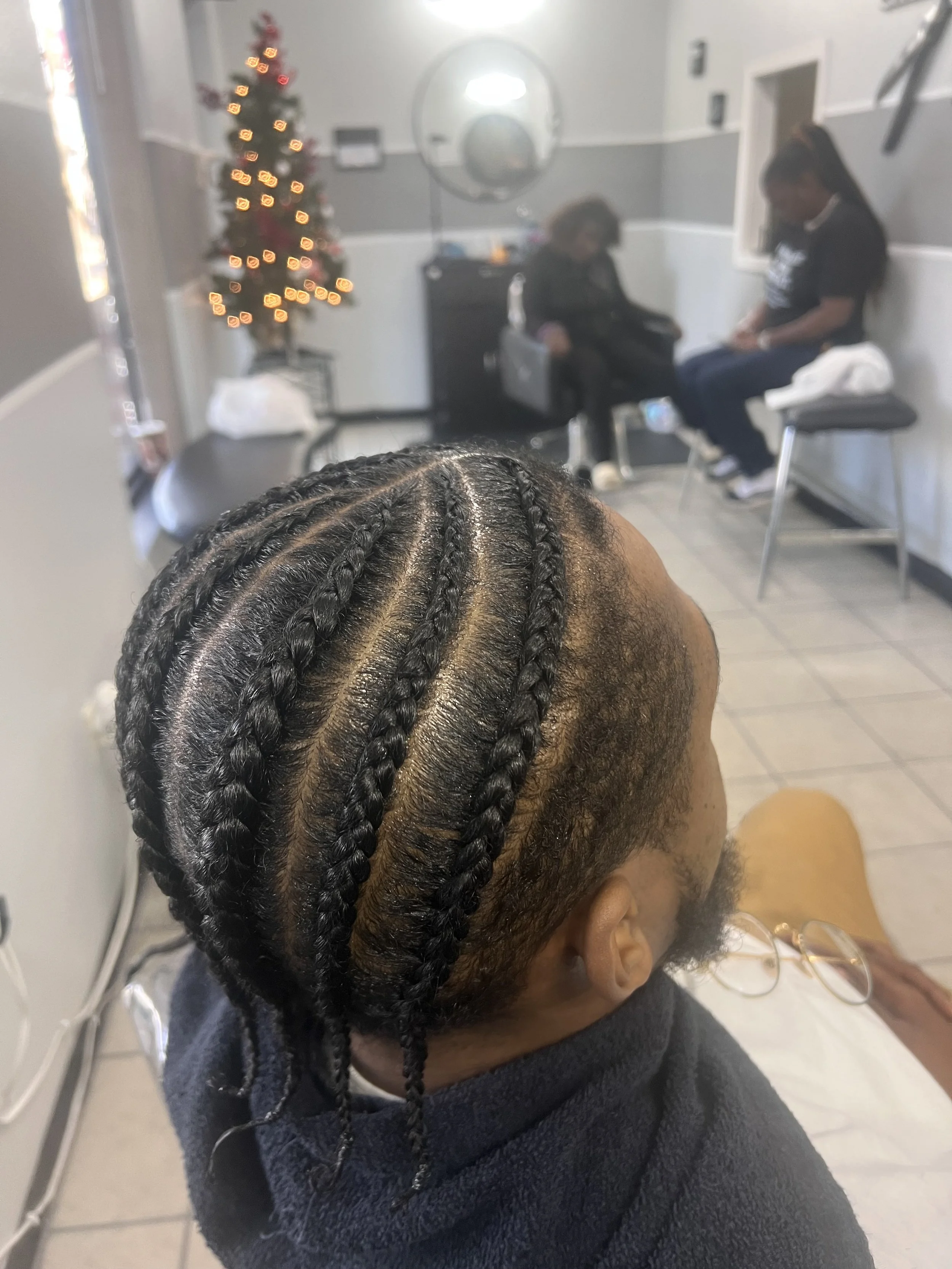 Person with freshly braided hair sitting in a salon, with a Christmas tree in the background and two women seated in chairs.