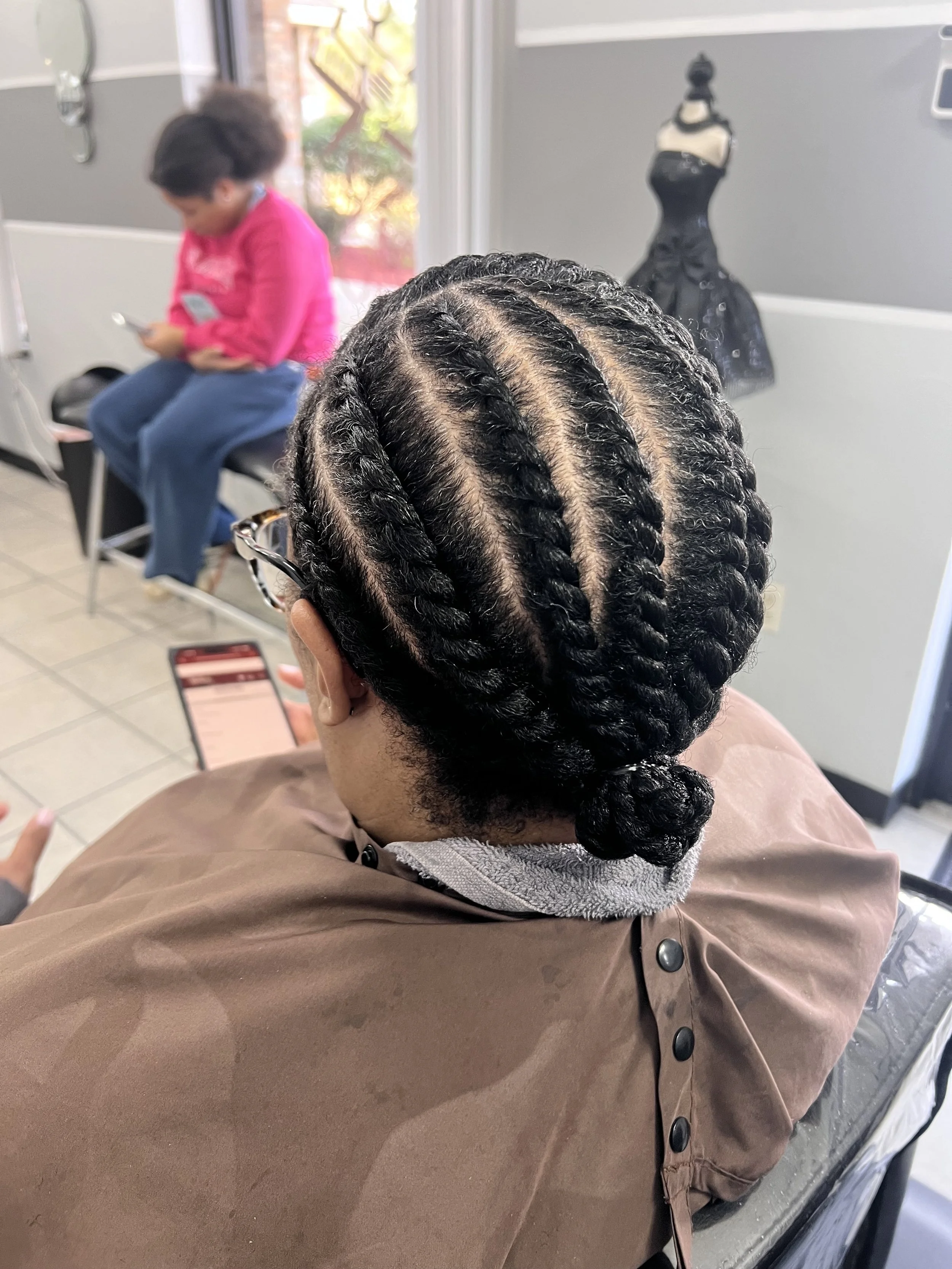 A person with freshly braided cornrow hairstyle sitting in a salon chair and looking at their phone. In the background, a woman in a pink top and blue jeans is sitting and using her phone, with a styling mannequin dressed in black behind her.