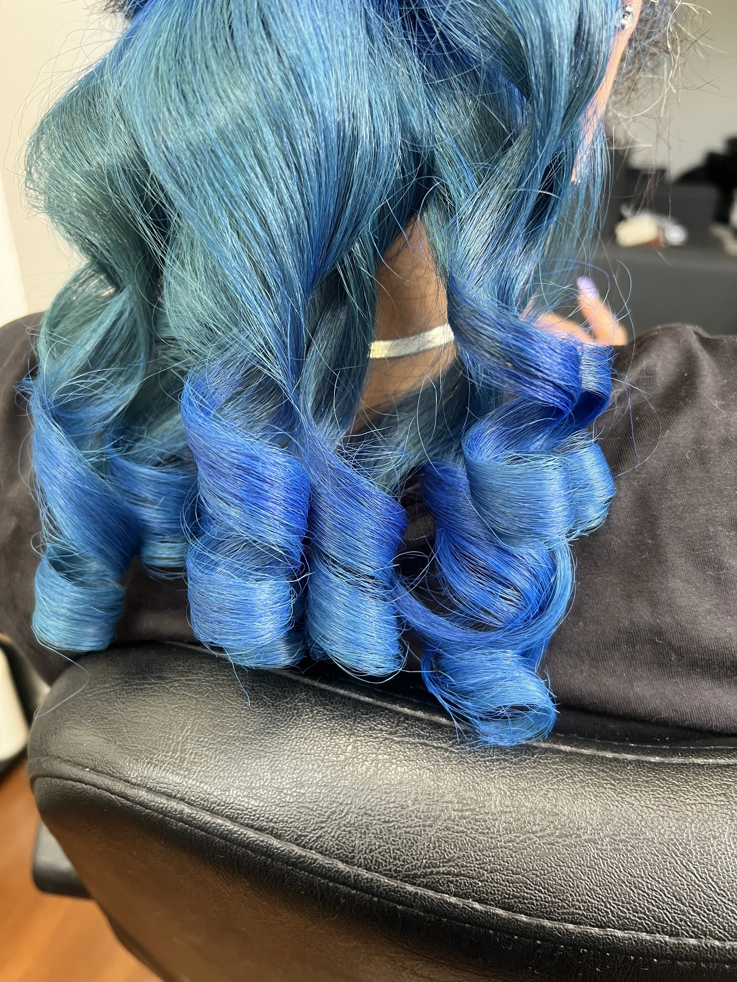 Close-up of a person's hair dyed in shades of blue, styled in loose curls, sitting on a black chair with a black salon cape.