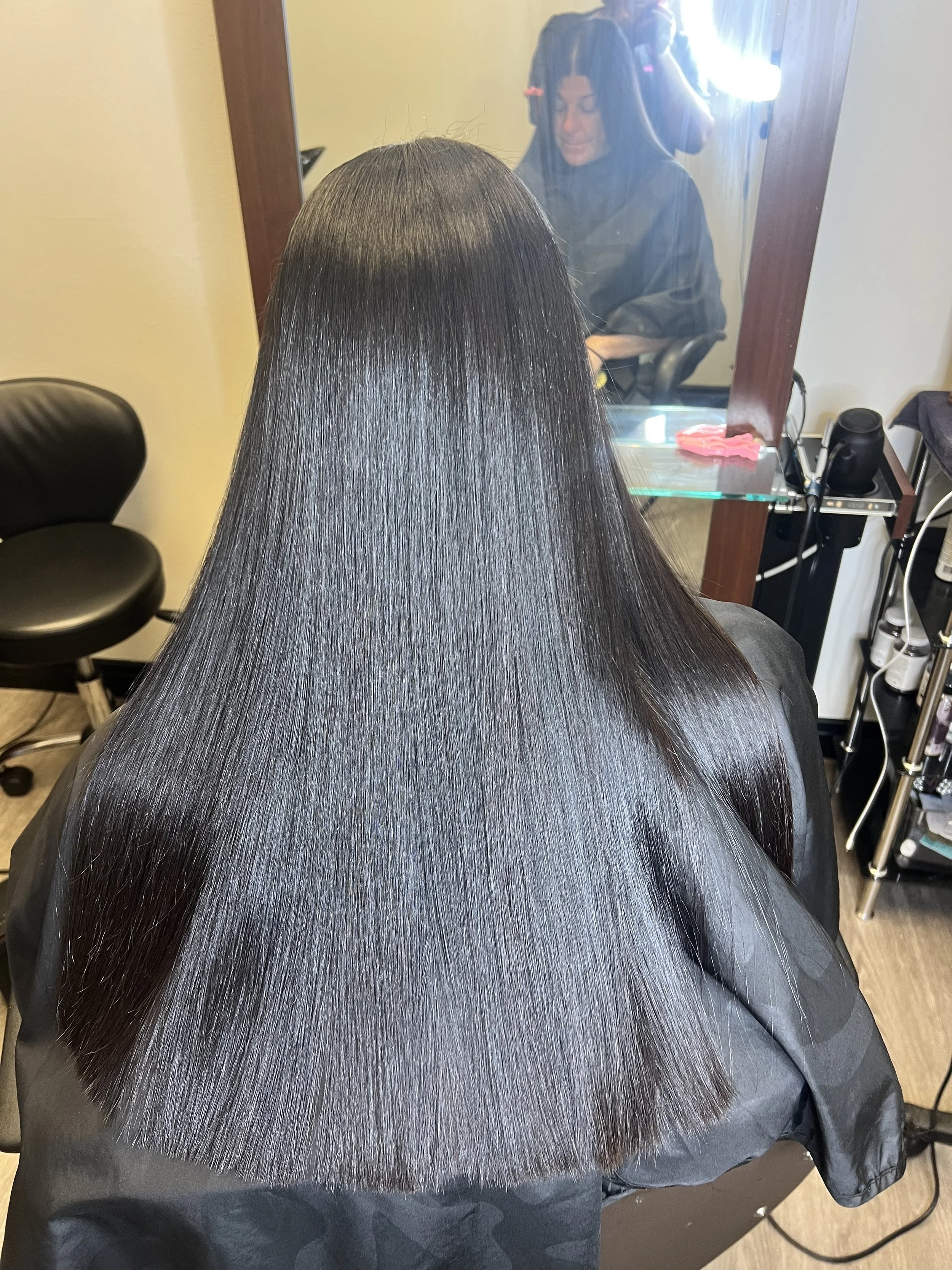 A woman with long, straight, shiny black hair sitting in a hair salon, with her reflection visible in a mirror behind her.