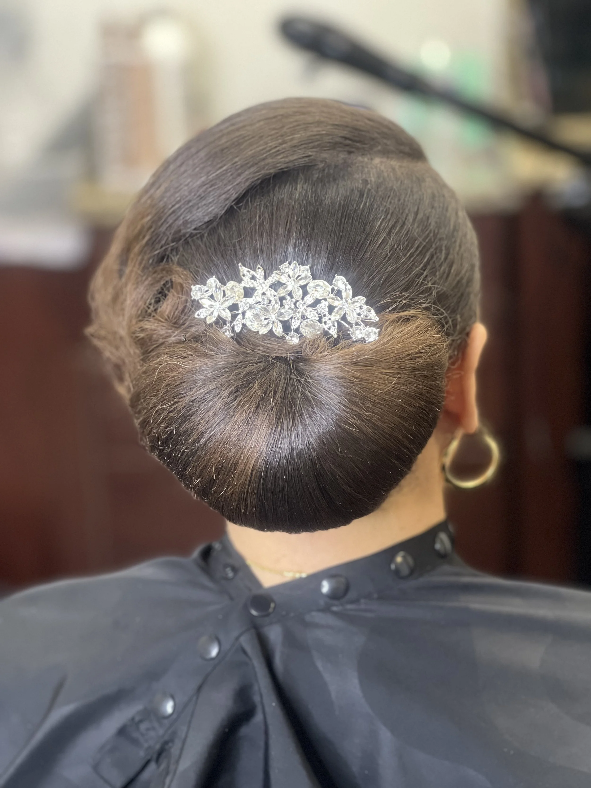 Up-close view of a woman's elegant hairstyle with a large rhinestone hair accessory, wearing a black cape with buttons, and gold hoop earrings.