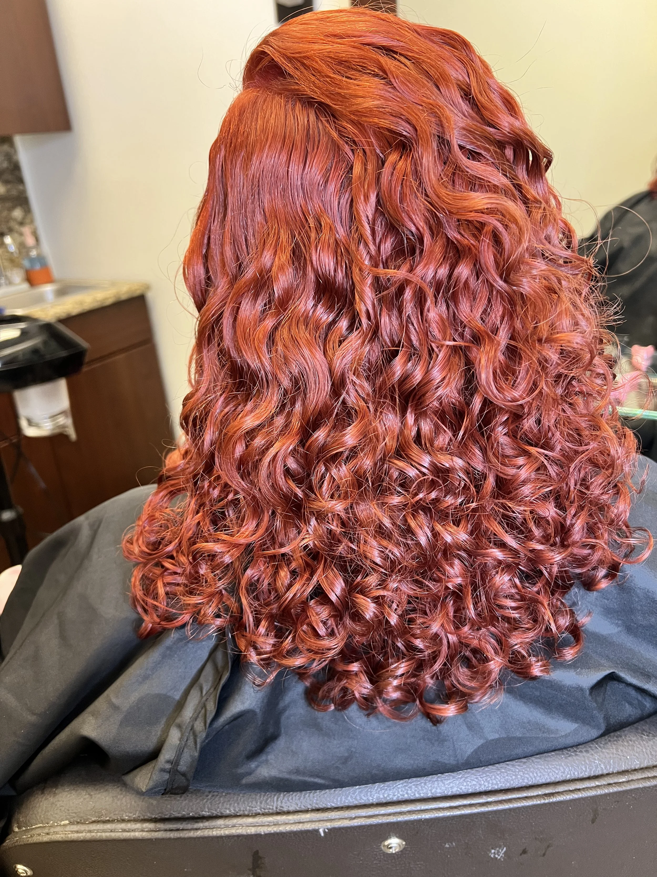 Back view of a person with vibrant red, curly hair styled in defined ringlets, sitting in a salon chair.