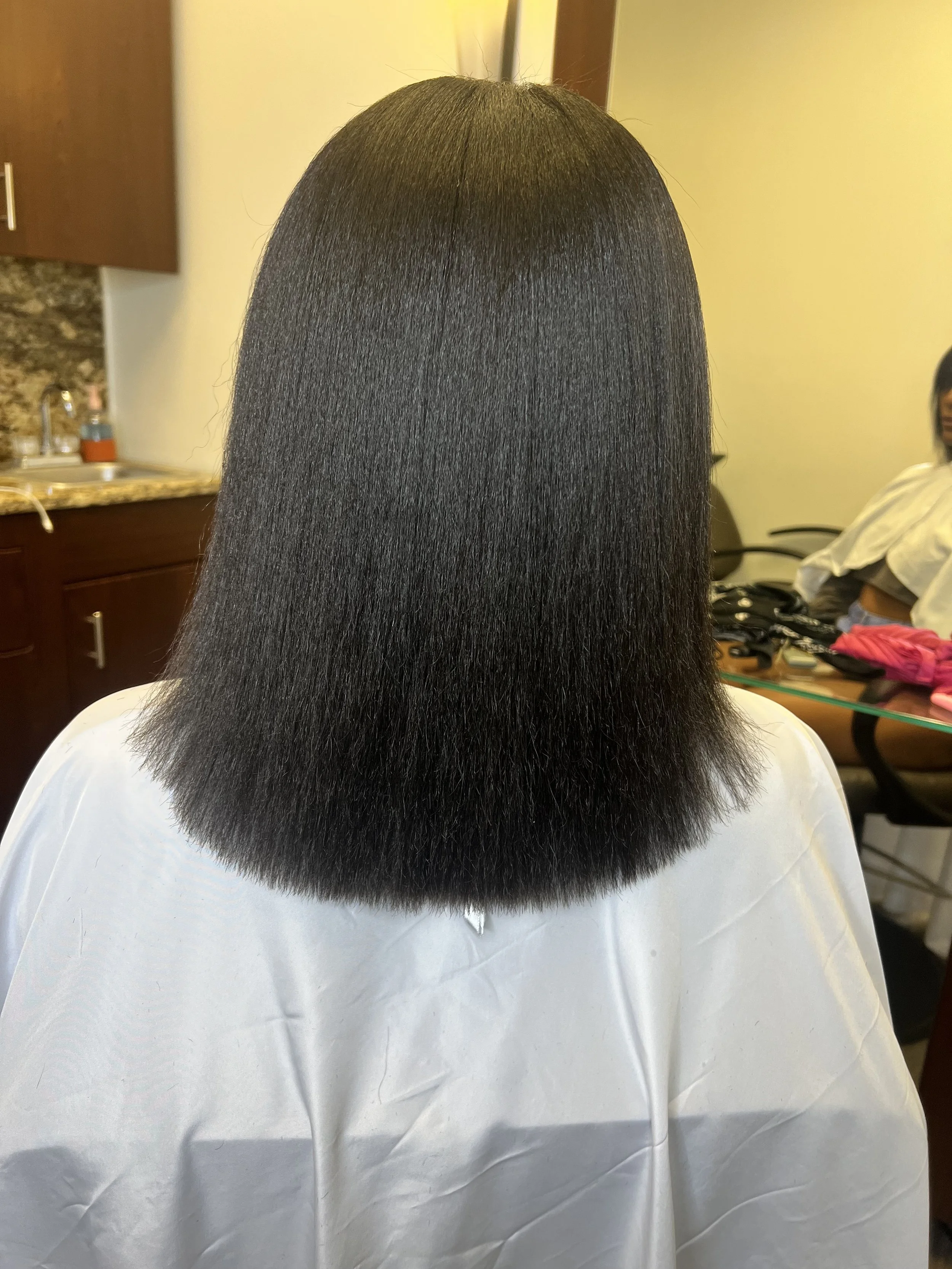 Person with straight, shiny black shoulder-length hair sitting in a salon chair wearing a white cape, with a salon background including another person, hair tools, and pink clothing.