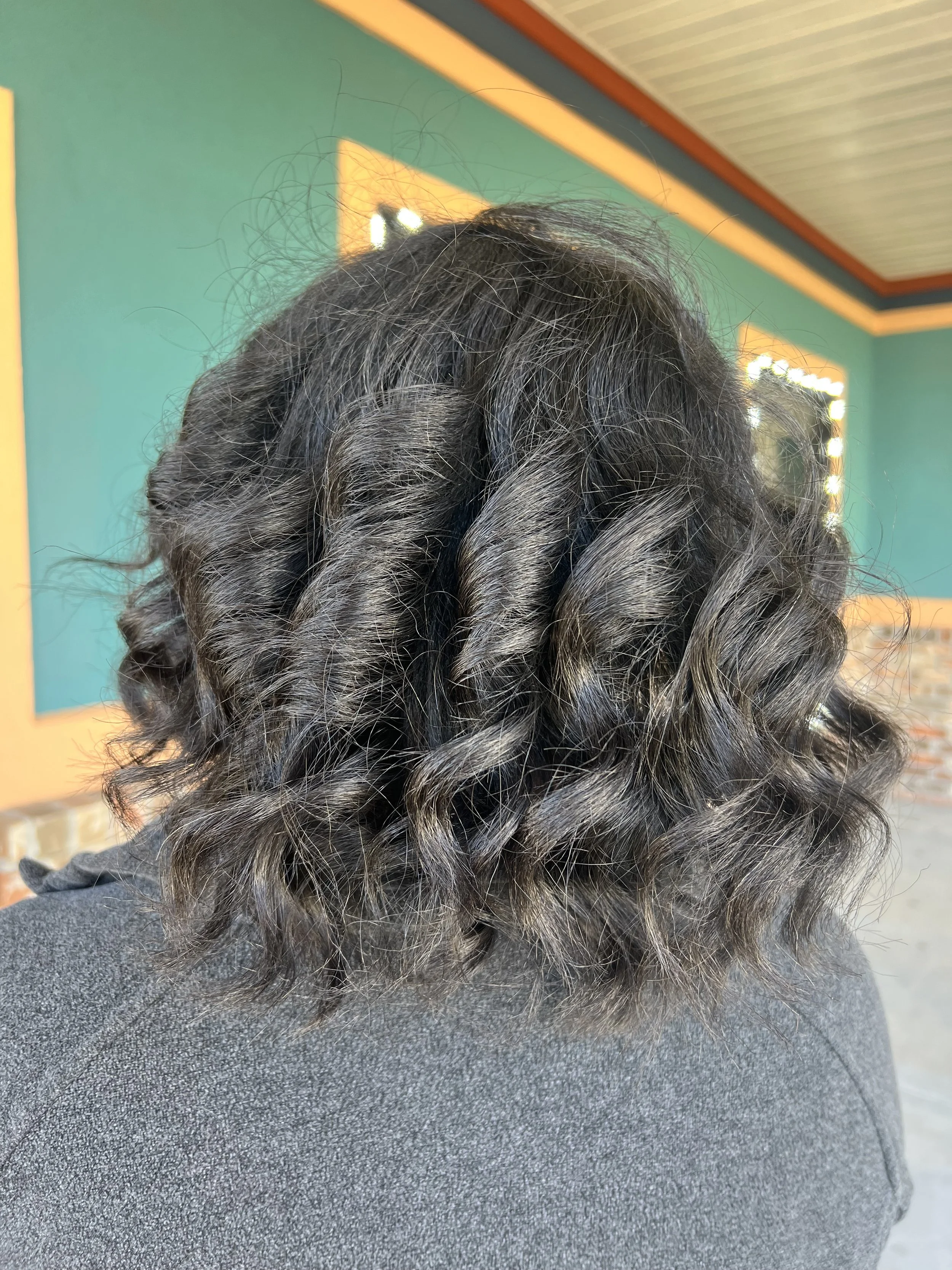 Back view of a person with shoulder-length dark curly hair, wearing a gray sweater, inside a colorful room with teal walls and orange trim.