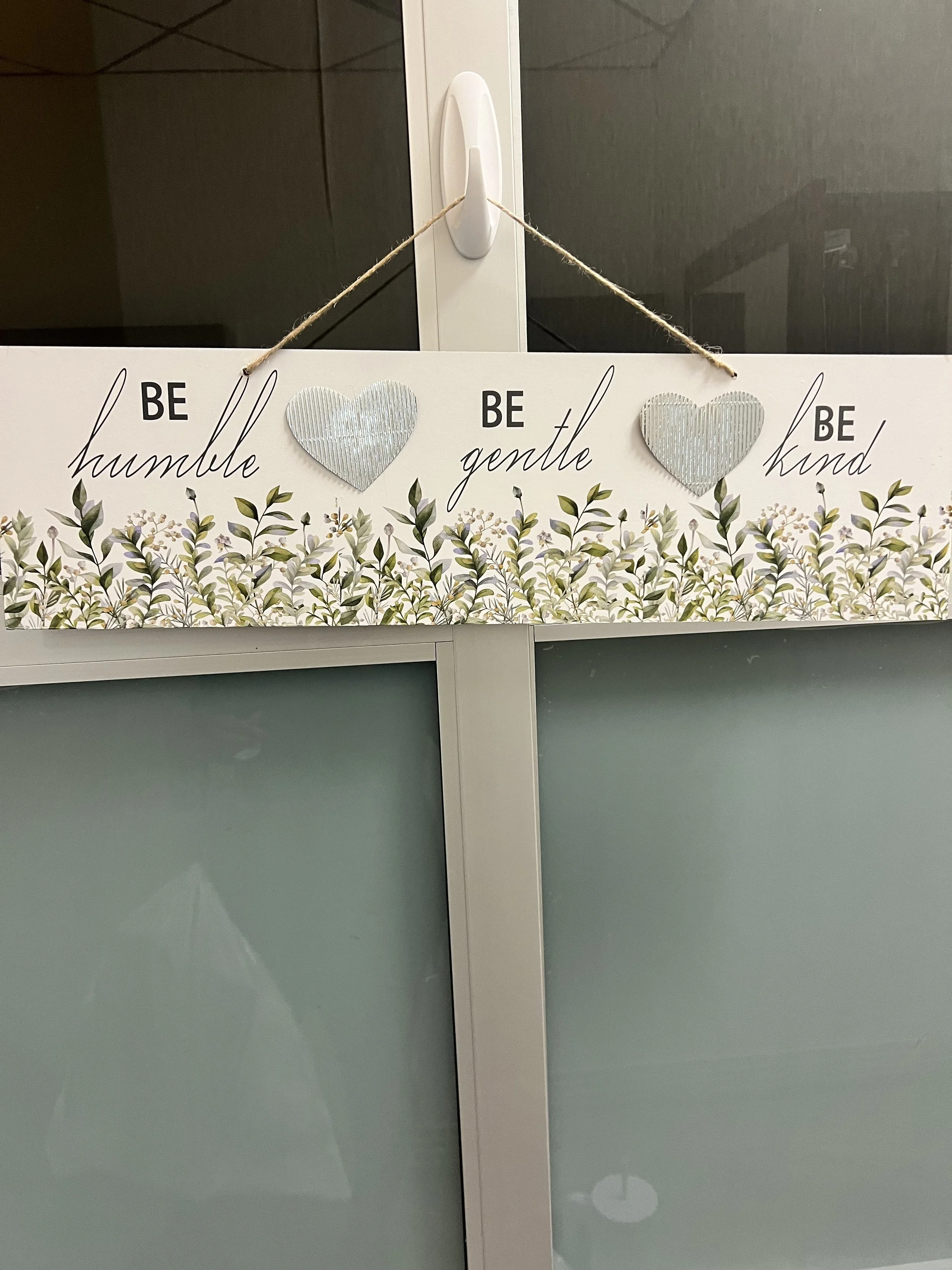 Decorative hanging sign with the words 'Be humble', 'Be gentle', and 'Be kind', featuring fabric heart shapes and a floral pattern at the bottom, attached to a glass door or window.