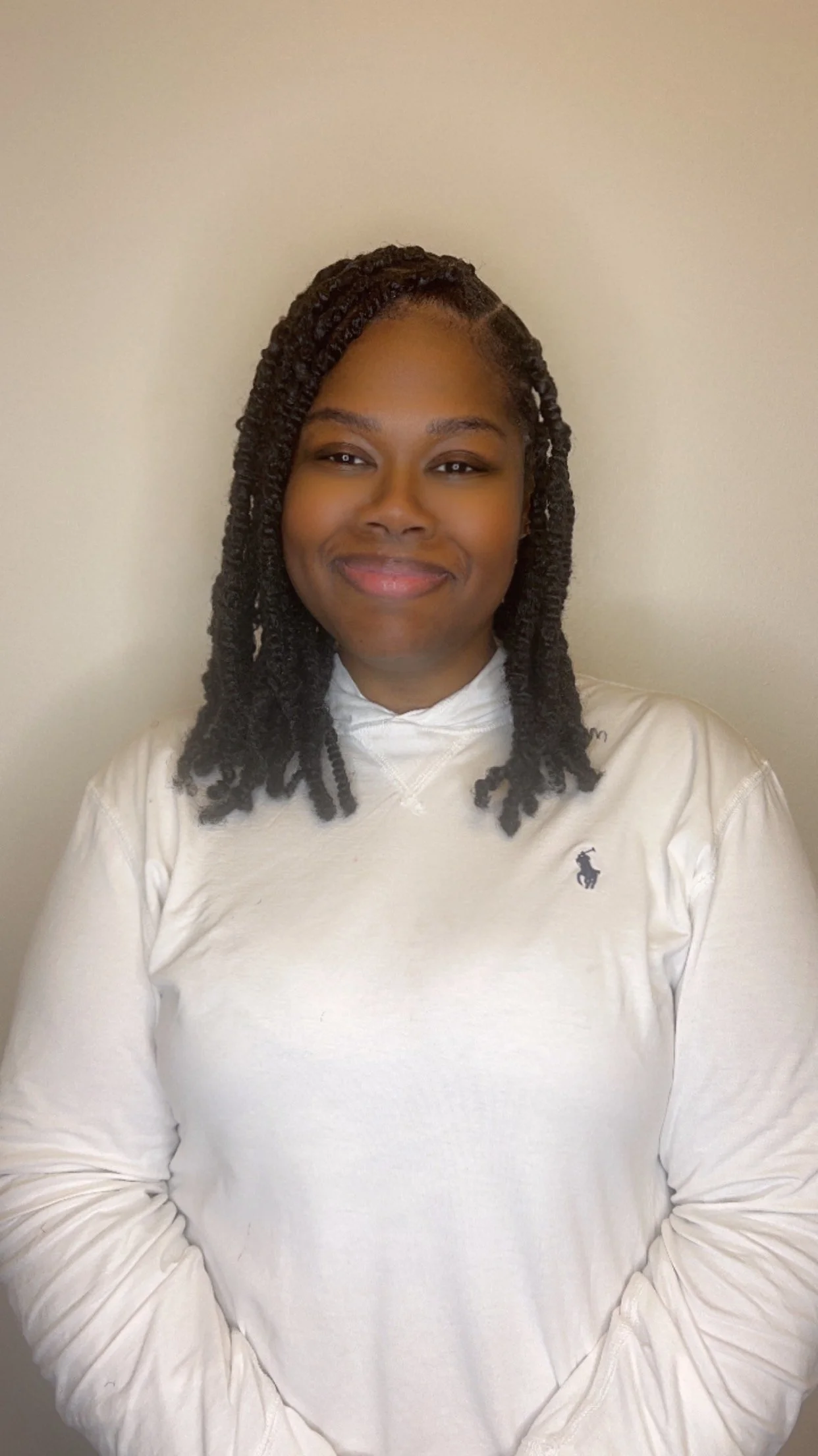 A smiling woman with dark braided hair, wearing a white long-sleeve shirt with a small logo on the chest, standing against a plain beige wall.