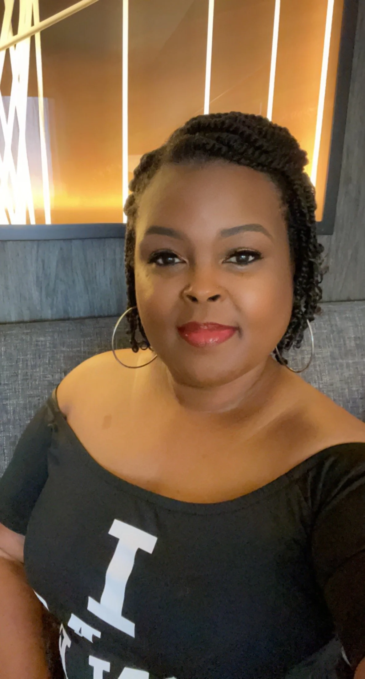 A woman with braided hair and hoop earrings smiling at the camera, wearing a black off-the-shoulder top with white text, seated against a dark textured wall with a decorative light feature in the background.
