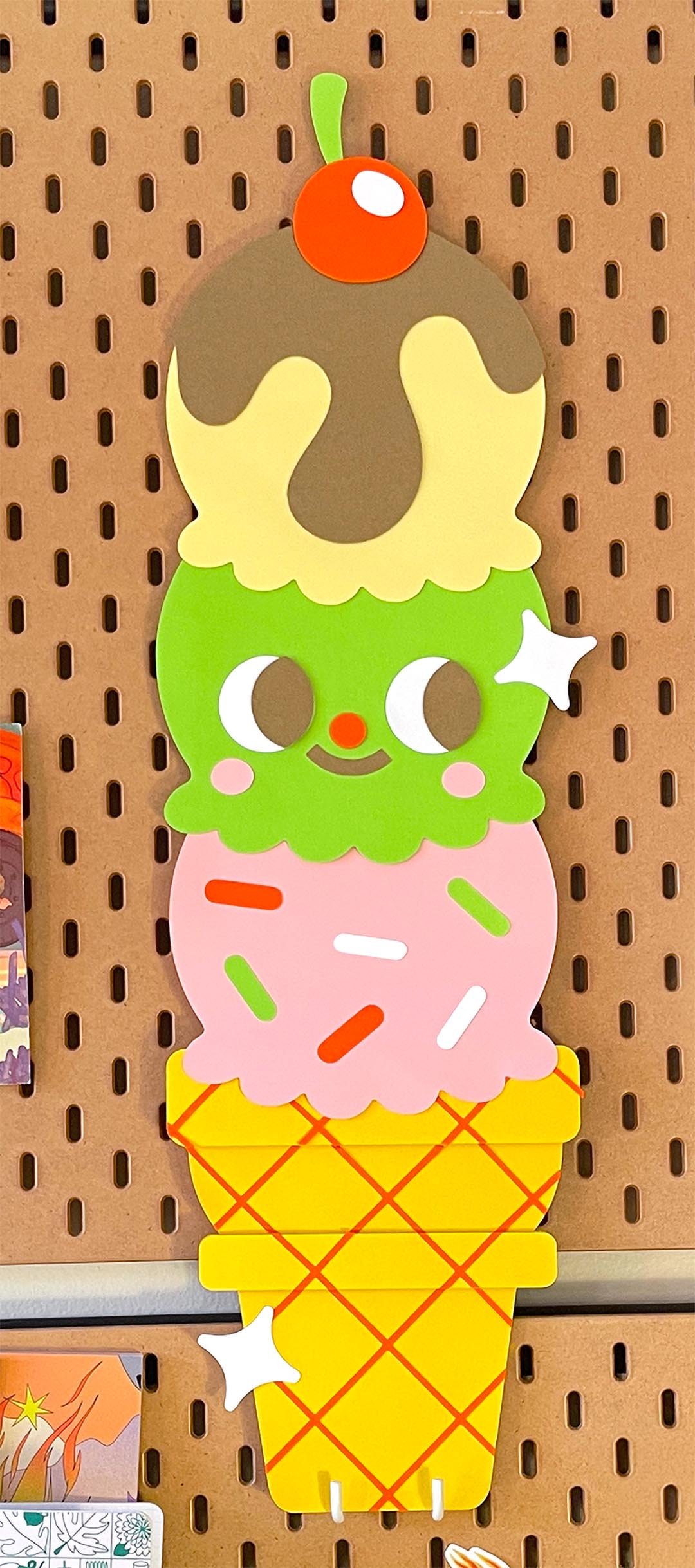 Colorful smiling ice cream cone character displayed on a pegboard in the Kidnichols studio. Constructed out of fine art paper.