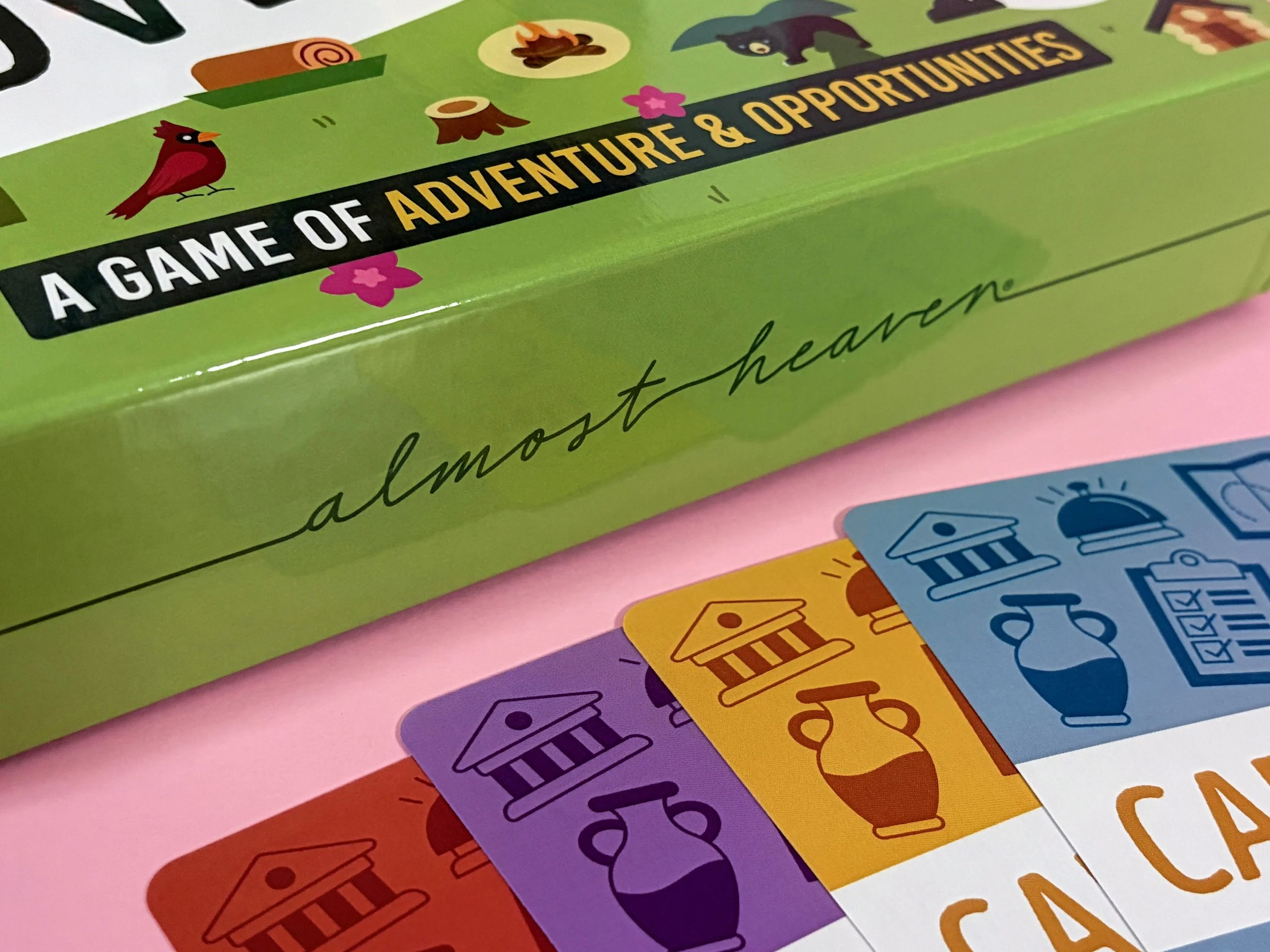 Close-up of a colorful board game box with the title "A Game of Adventure & Opportunities" and game cards featuring various illustrations and colors.