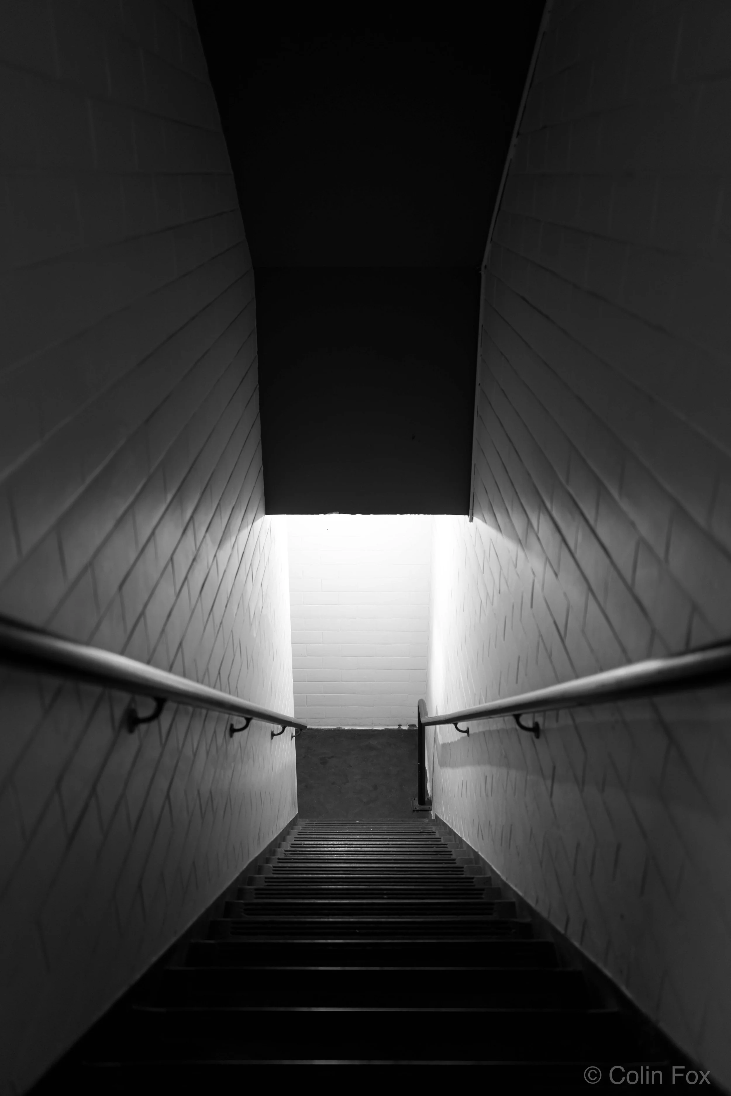 Black and white photo of a downward staircase with light at the bottom, metal handrails on both sides, enclosed by textured wall panels.