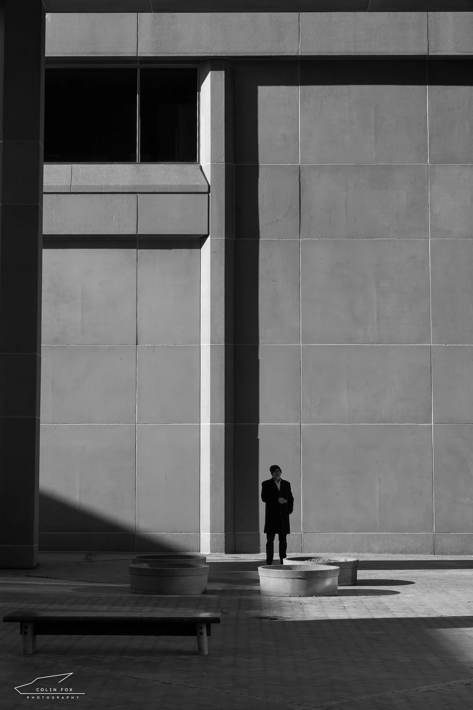 A man in a coat standing outdoors in front of a large building wall with a shadow cast across it. There are round concrete planters and a bench nearby, all in black and white.
