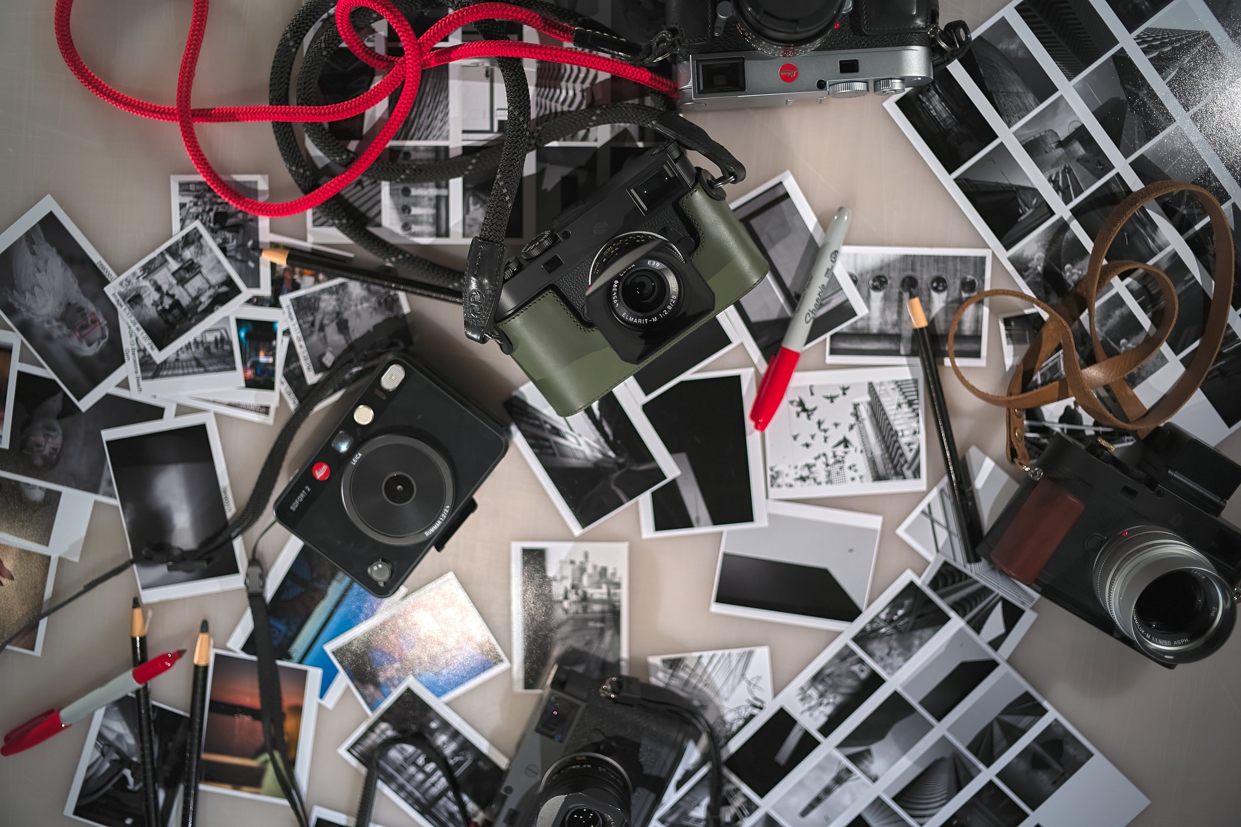 A cluttered table with black-and-white photographs, two vintage cameras, a flash, a red and black marker, and scattered camera straps and cables.