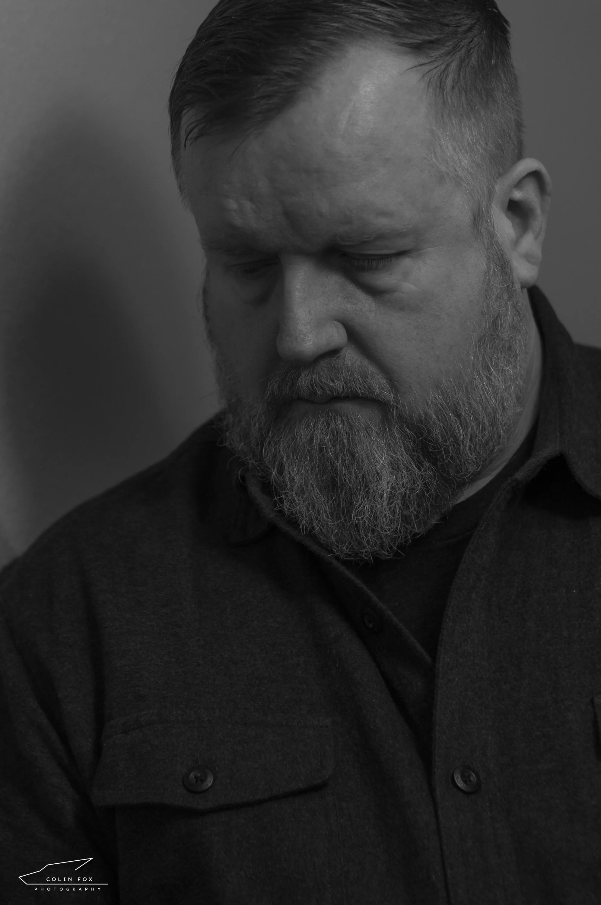 Black and white portrait of a man with a beard and short hair, looking down, wearing a dark button-up shirt.