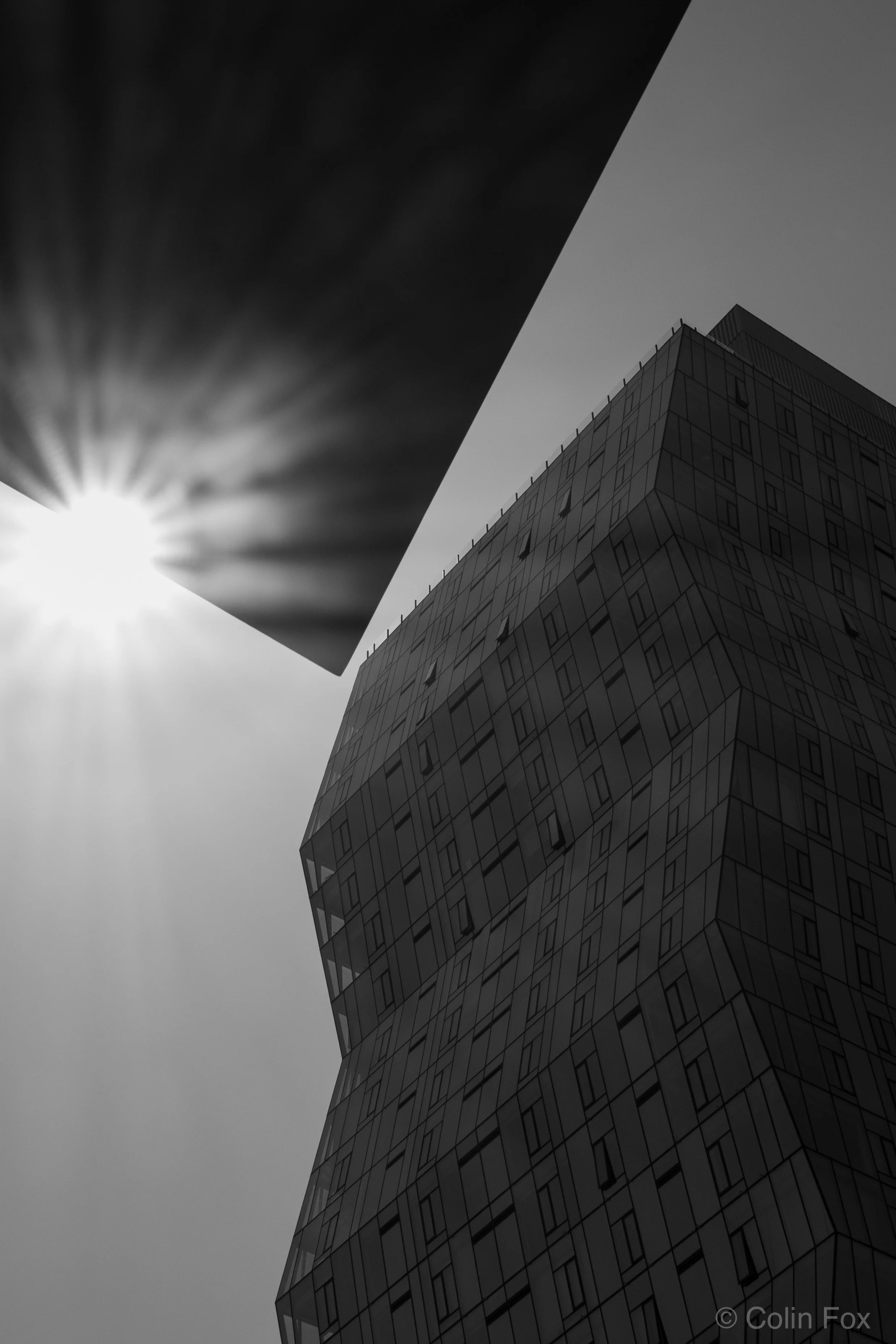 A black and white photo of a modern, angular glass building with a geometric design, set against a clear sky with the sun shining brightly in the background.