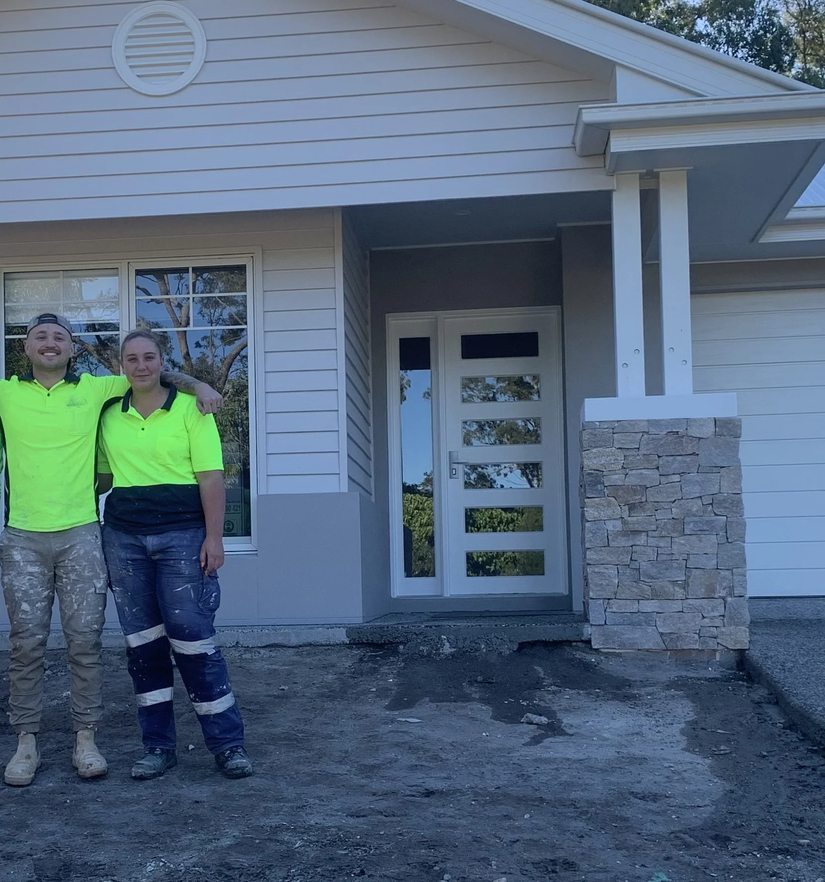 Two construction workers in neon yellow shirts stand in front of a house with a modern front door and stone accents, smiling at the camera.