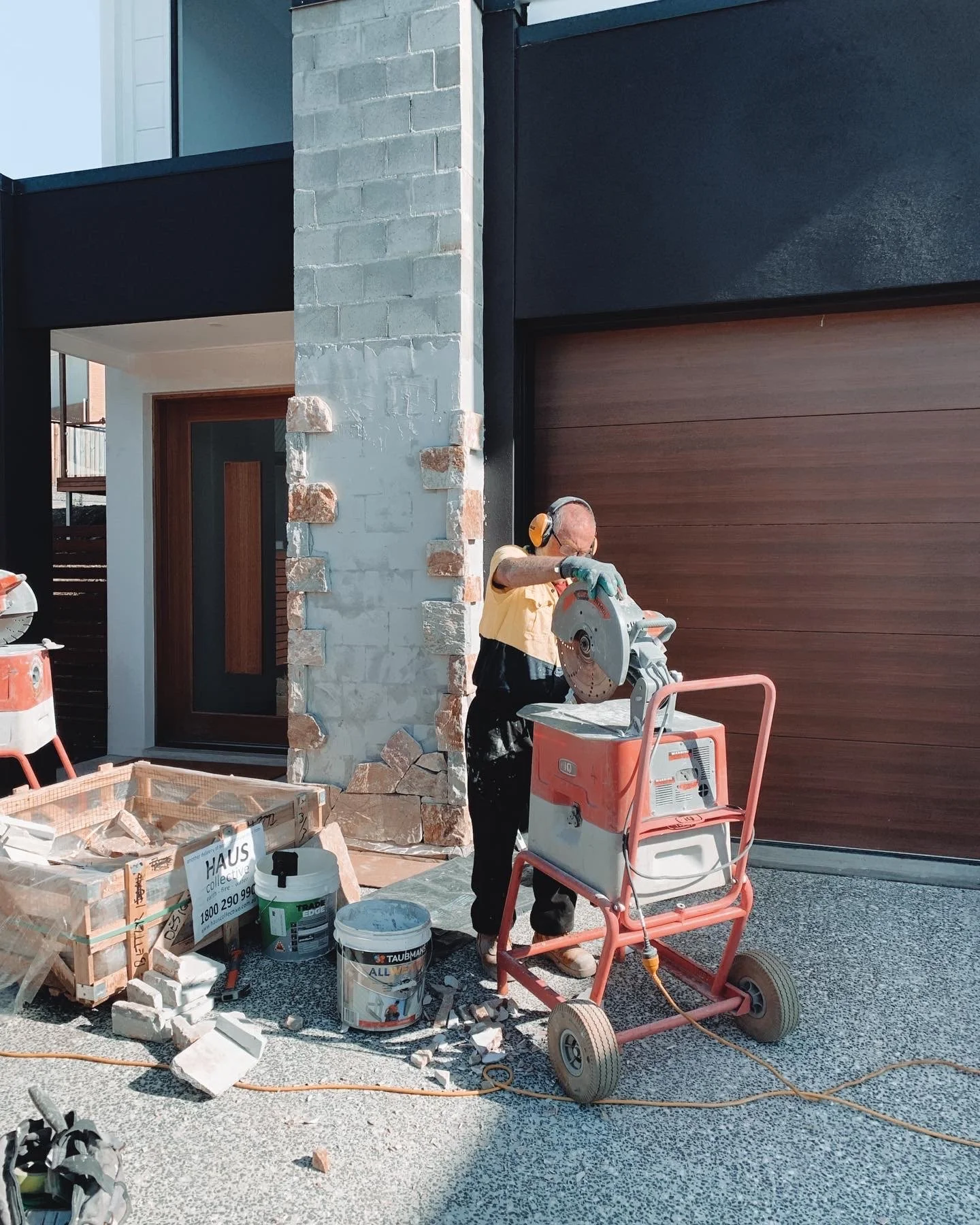 A man wearing yellow shirt, black pants, and gloves uses a circular saw to cut a piece of stone or concrete next to a house with a stone and wood exterior. There are construction supplies and tools on the ground, including a bucket, a cardboard box, and a work cart.