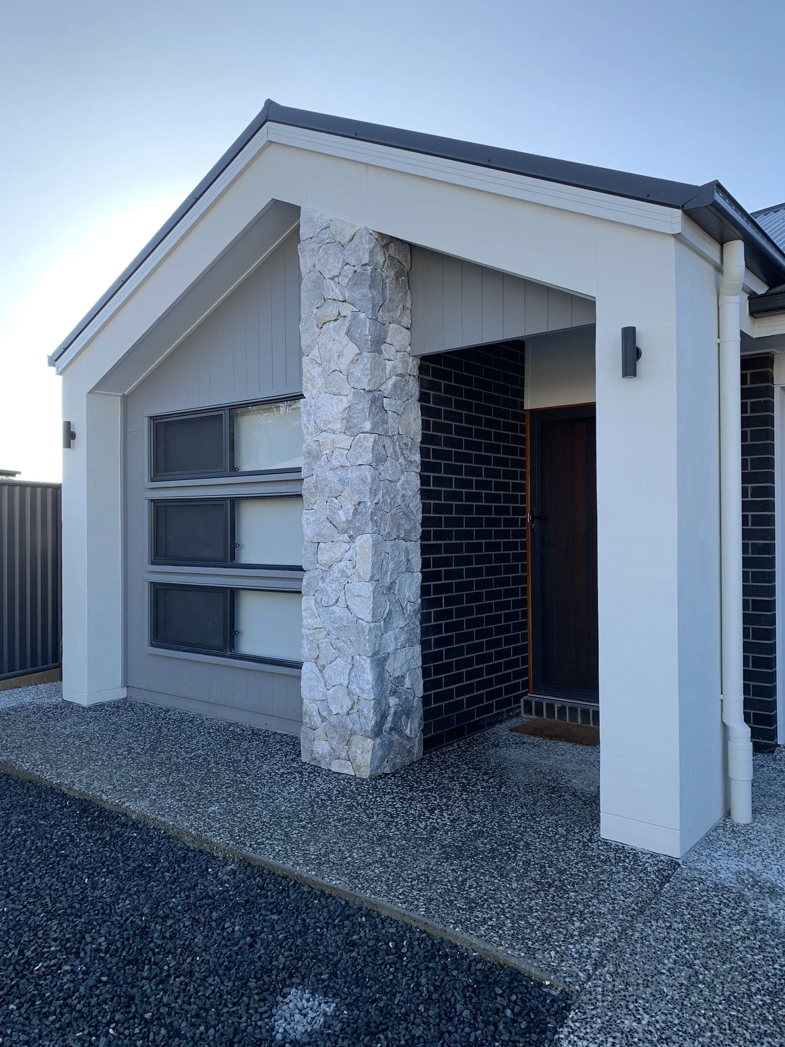 Modern house entrance with a black wooden door, black brick wall, stone column, and a large area of concrete paving.