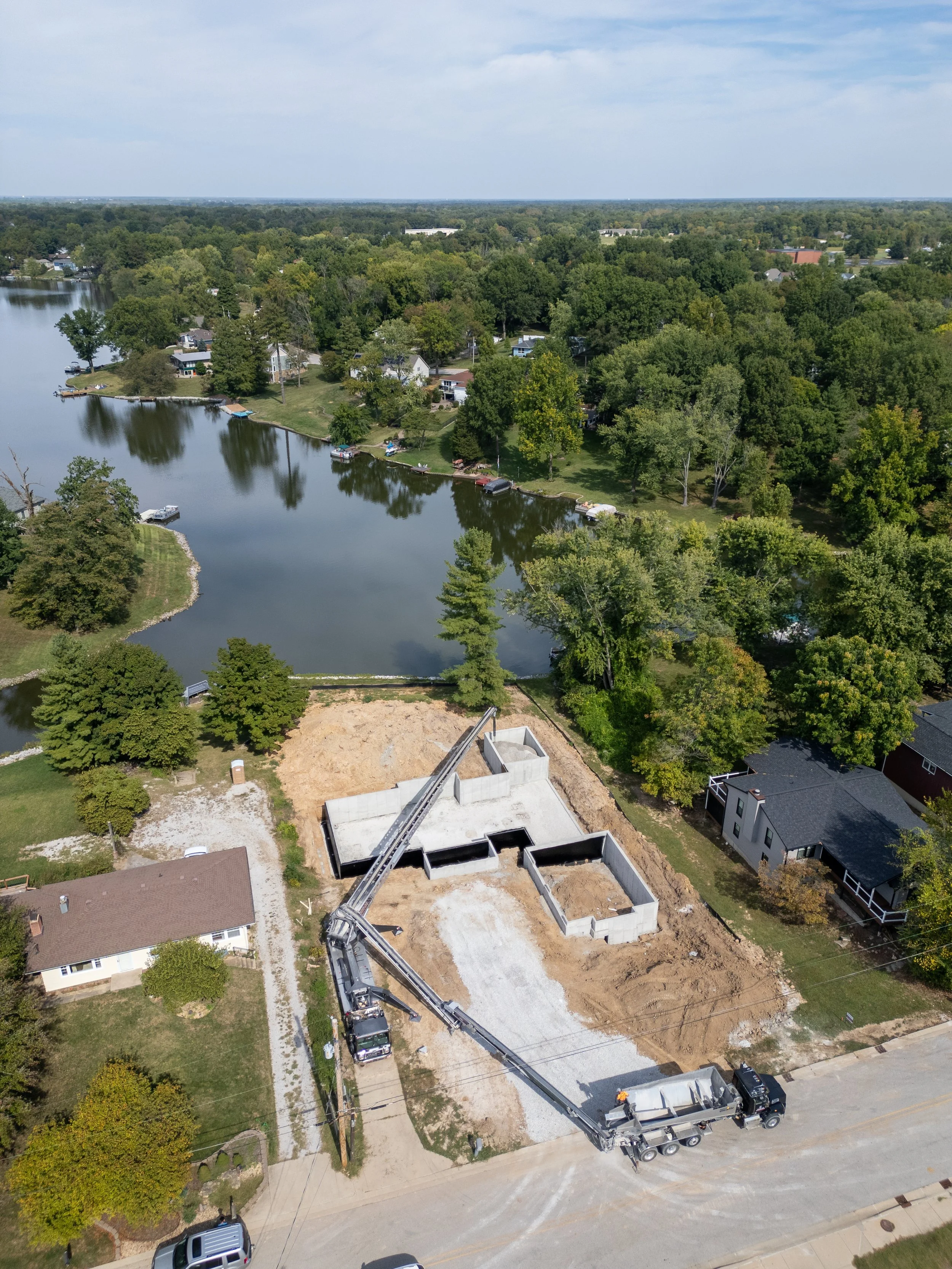 An aerial view of a construction site with a partially built house and a large crane. The site is in a suburban neighborhood next to a lake, with trees and houses surrounding the area.