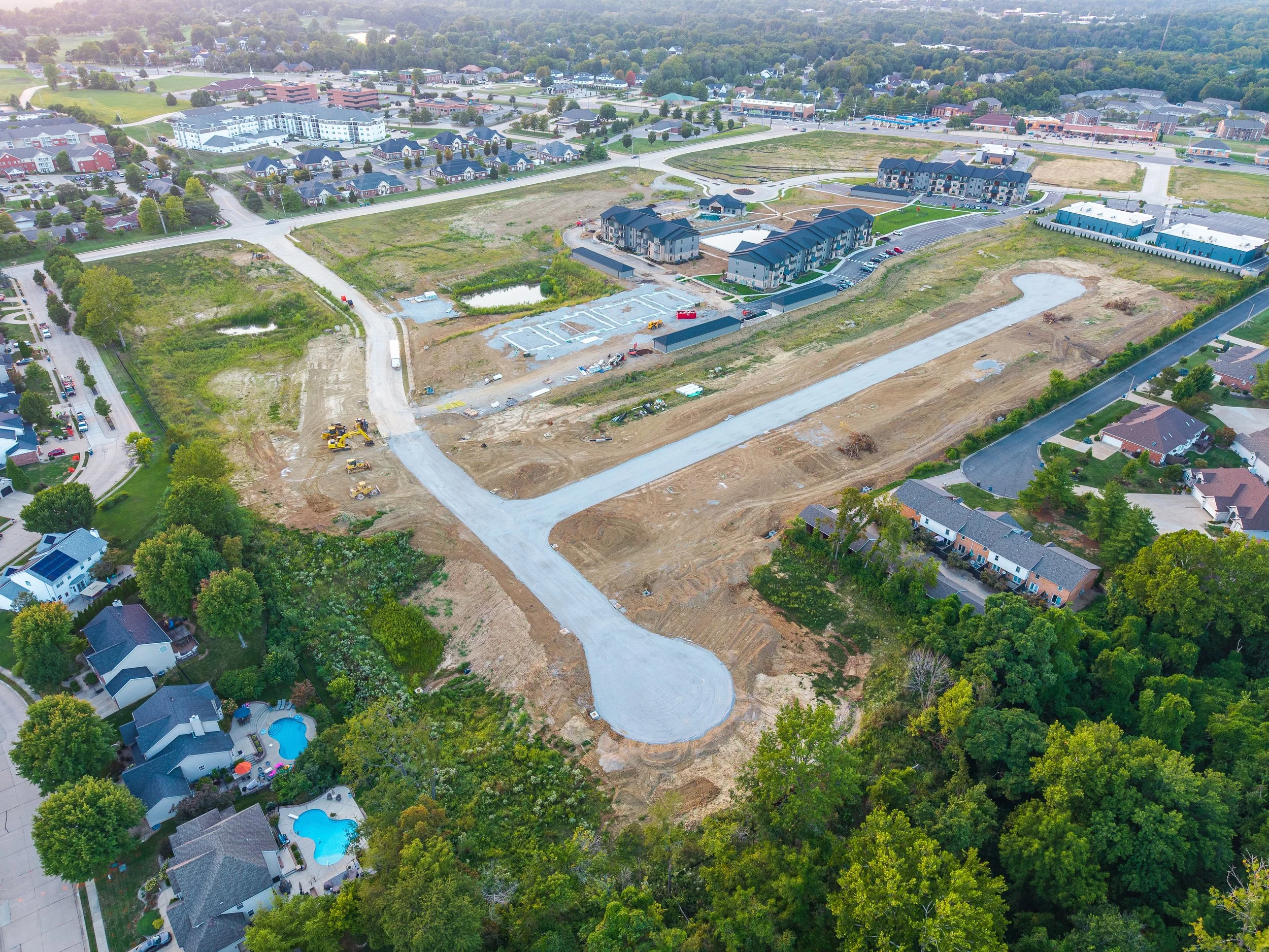 Aerial view of a construction site with new roads being built, surrounded by residential houses, apartment buildings, and green trees.
