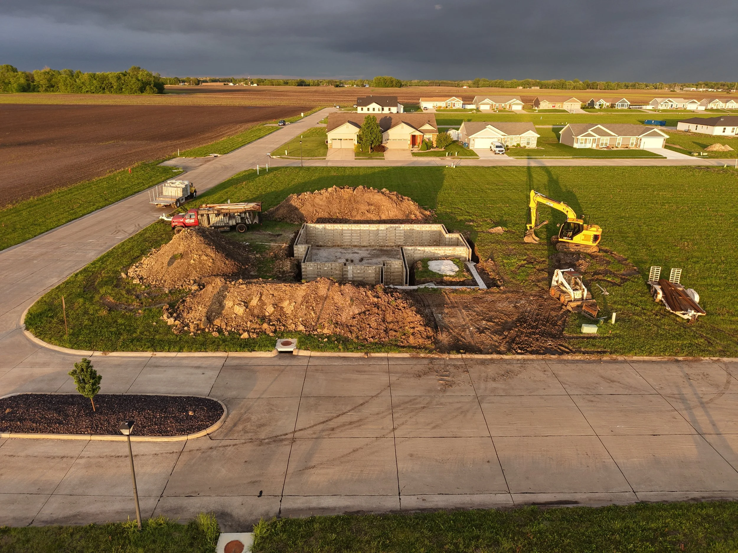 Construction site in a suburban neighborhood with an excavator, small trucks, and a foundation being built, with residential homes and open fields in the background under a cloudy sky.